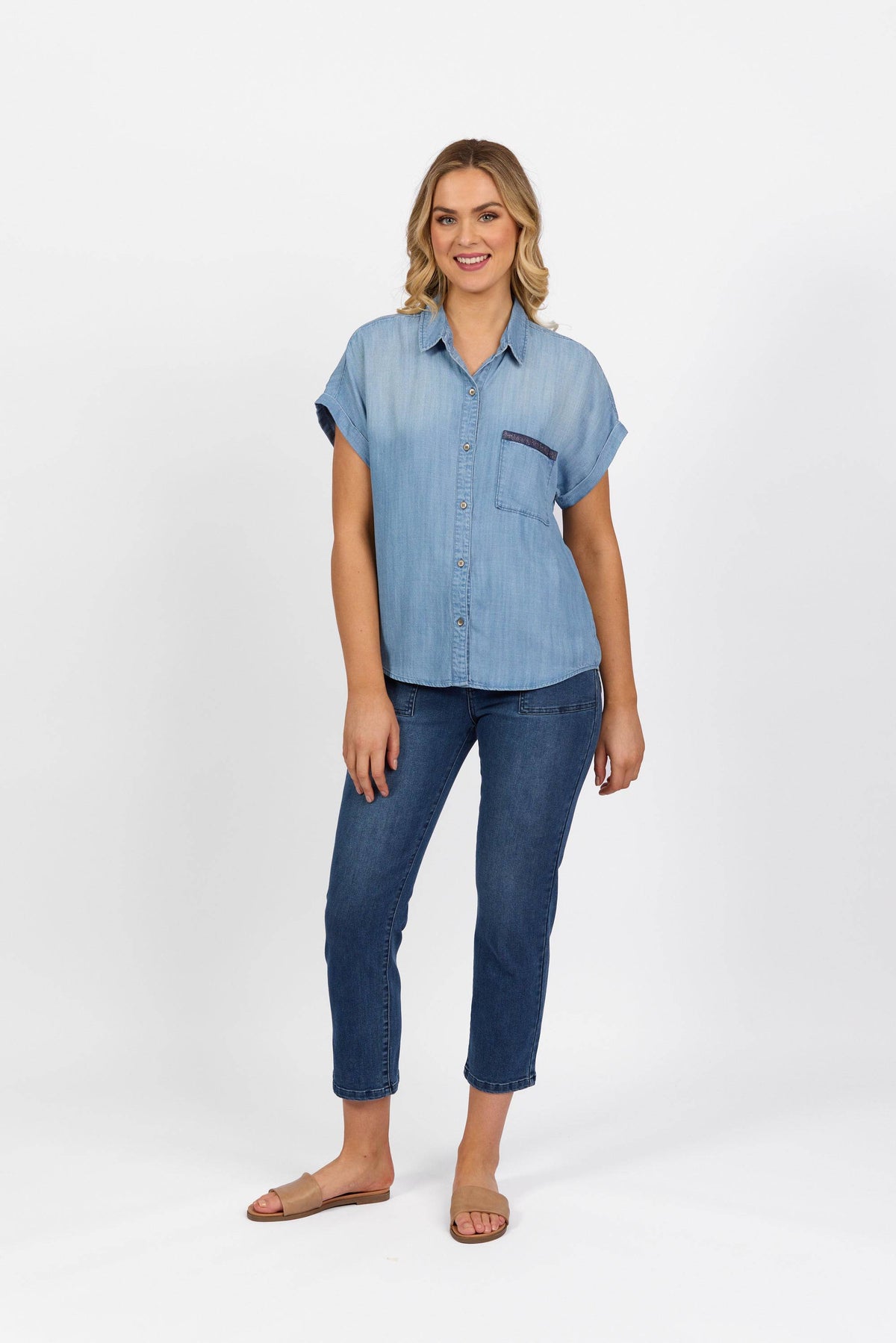 A woman stands against a white background, smiling and relaxed, wearing the Vassalli Shirt - Blue Wash with short sleeves and dropped shoulders, paired with blue jeans and tan slide sandals.