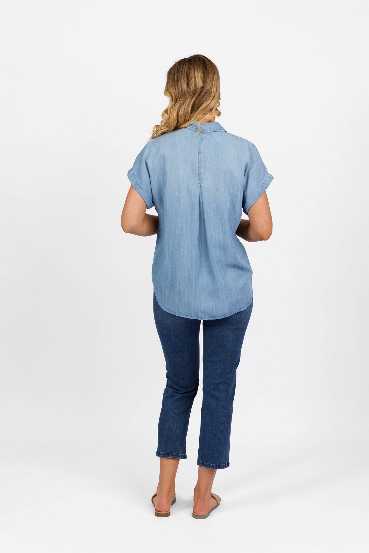 A woman stands facing away from the camera, wearing the Vassalli Shirt - Blue Wash, paired with dark blue jeans and sandals, her wavy blonde hair cascading down her back.