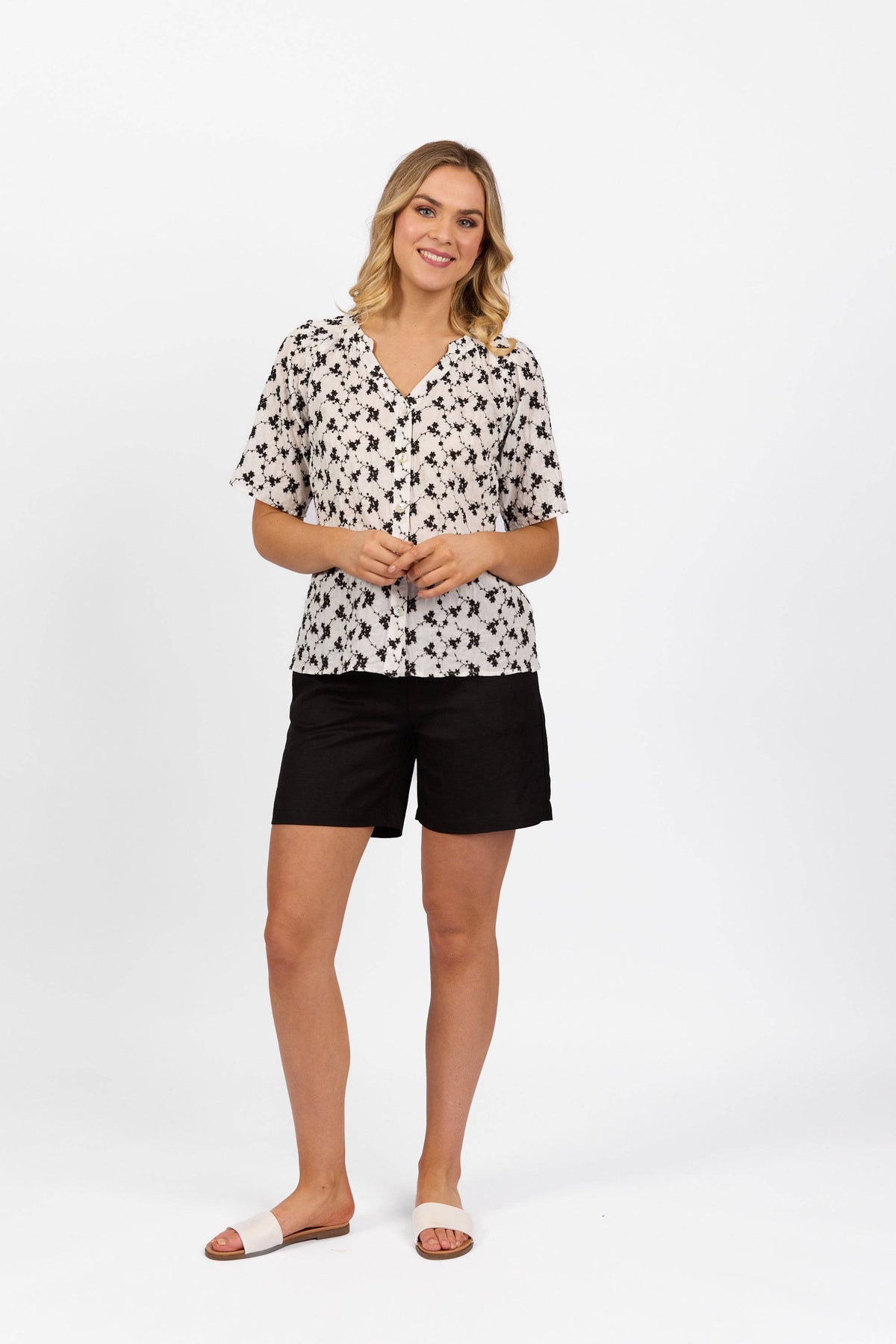 A woman smiles against a white background, wearing the Vassalli V-Neck Top - Floral, paired with black shorts and white sandals. Her hair is in loose waves and her hands are gently clasped in front.