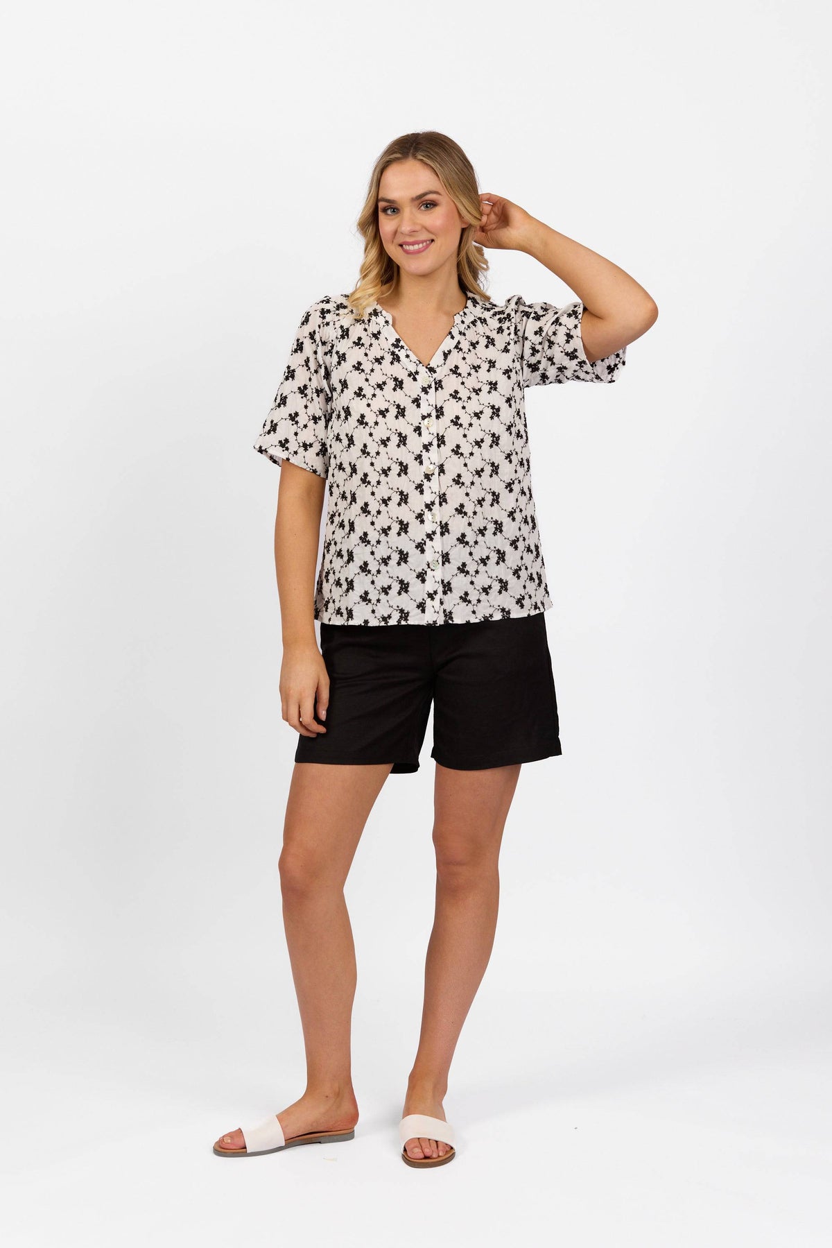 A woman stands against a white background, smiling and touching her hair. She wears the Vassalli V-Neck Top – Floral, paired with black shorts and white slide sandals.