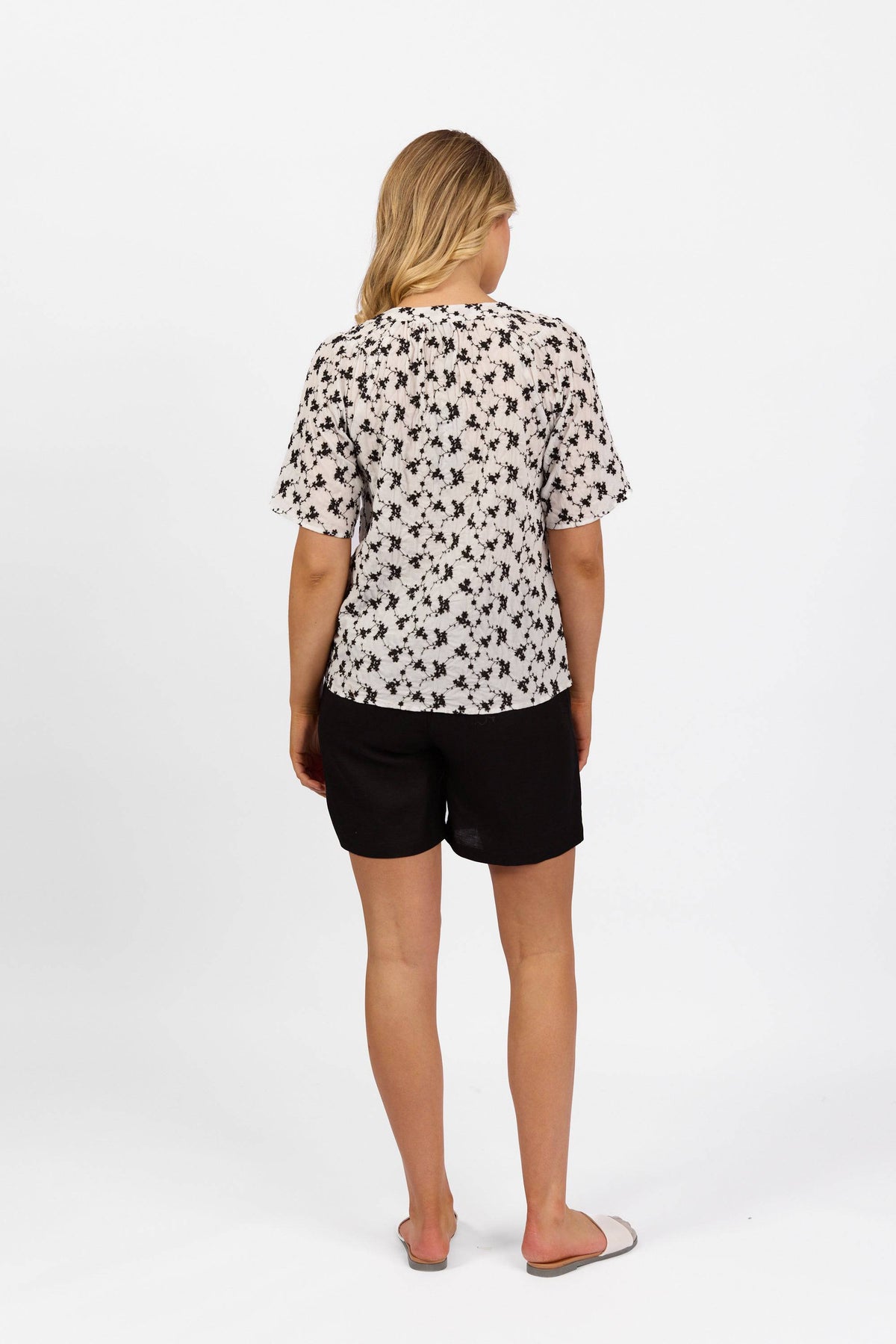 A woman faces away from the camera, wearing the Vassalli V-Neck Top - Floral, paired with black shorts and flat sandals, set against a plain white background.