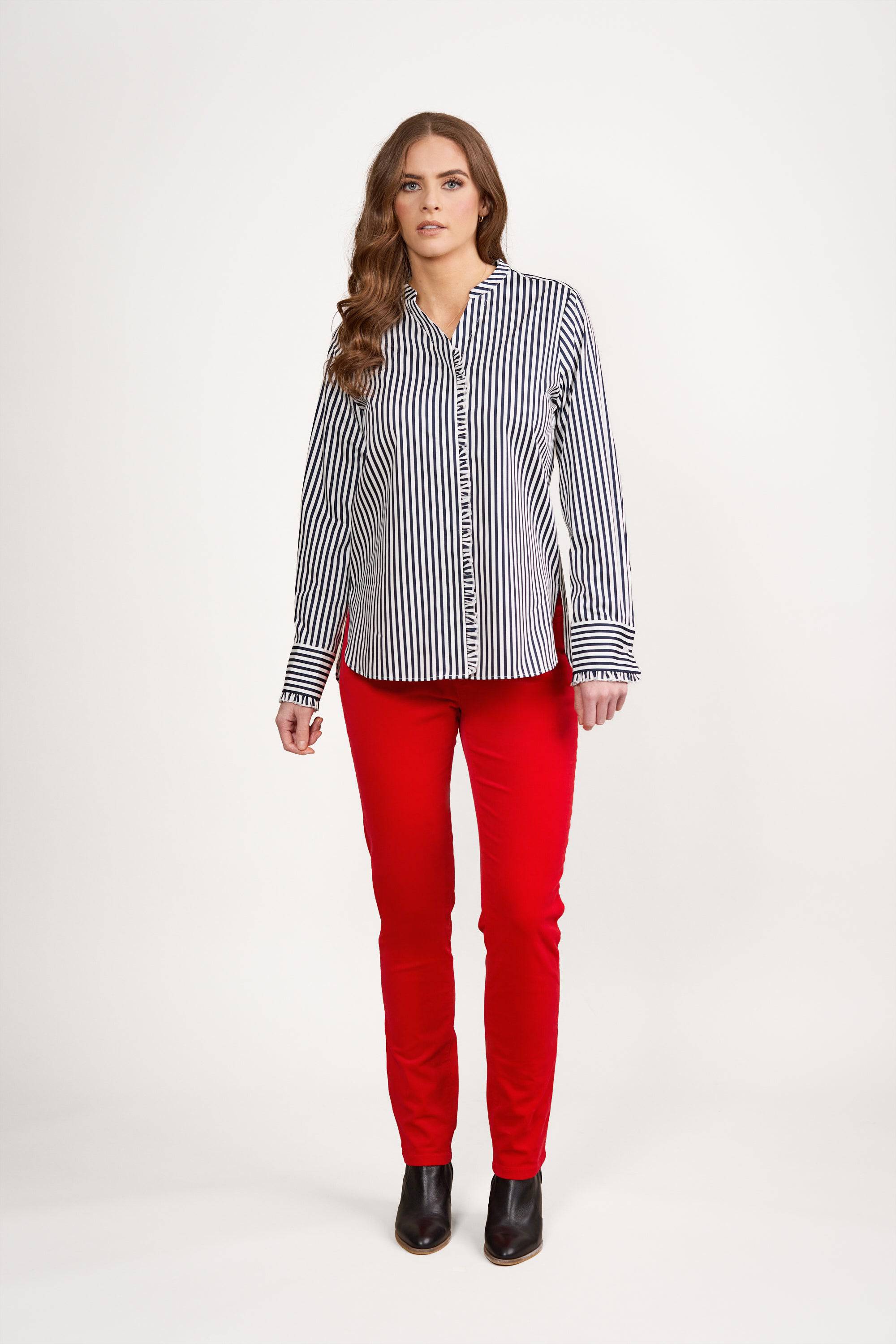 A woman stands facing forward against a white background, wearing the Vassalli Manhattan Shirt in Ink/White with vertical stripes and ruffle trim, bright red pants, and black ankle boots.
