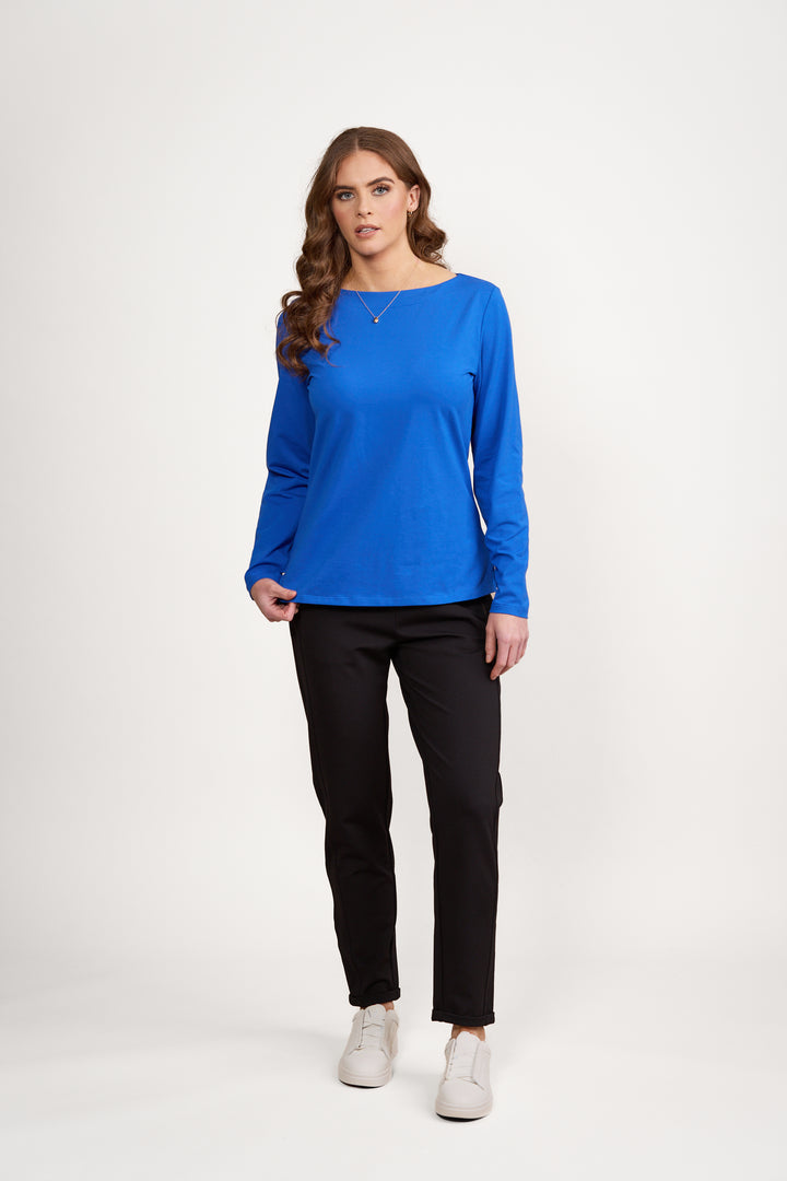 A woman with long, wavy brown hair stands against a white background, wearing the Vassalli Boat Neck Top in Electric Blue with black pants and white sneakers. She faces forward with a neutral expression.