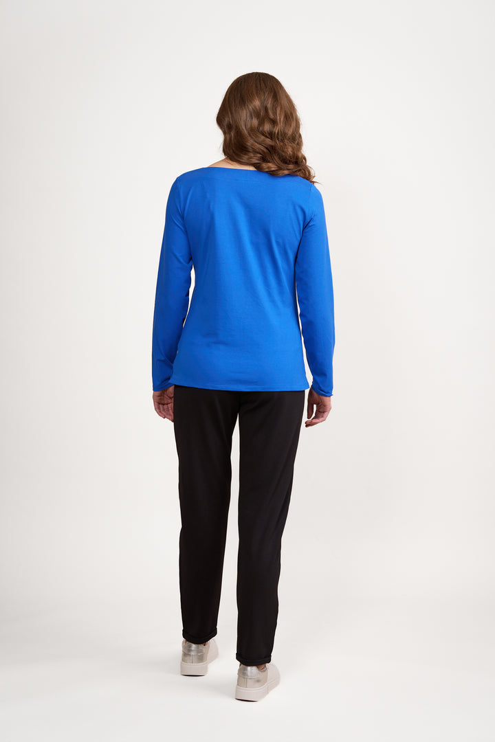 A person with wavy brown hair faces away, wearing the Vassalli Boat Neck Top in Electric Blue, paired with slim fit black pants and beige shoes, set against a plain white background.