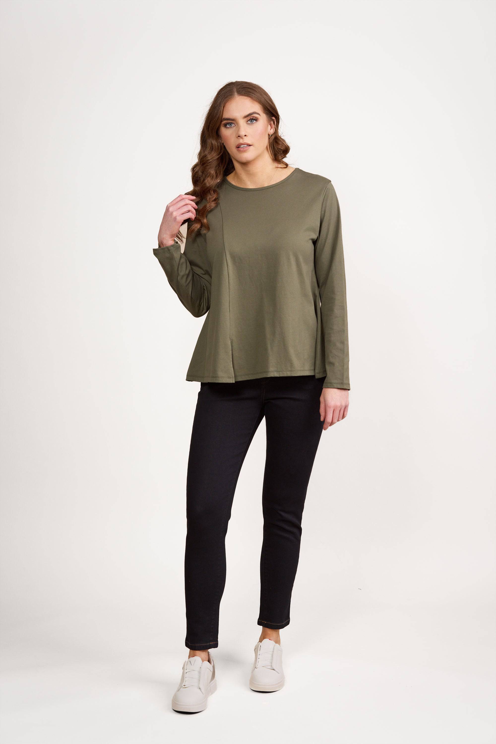 A woman with long brown hair wears the Vassalli Swing Top in Sage, featuring a relaxed fit and round neck. She pairs it with black pants and white sneakers, standing against a plain white background, glancing to the side with one hand in her hair.
