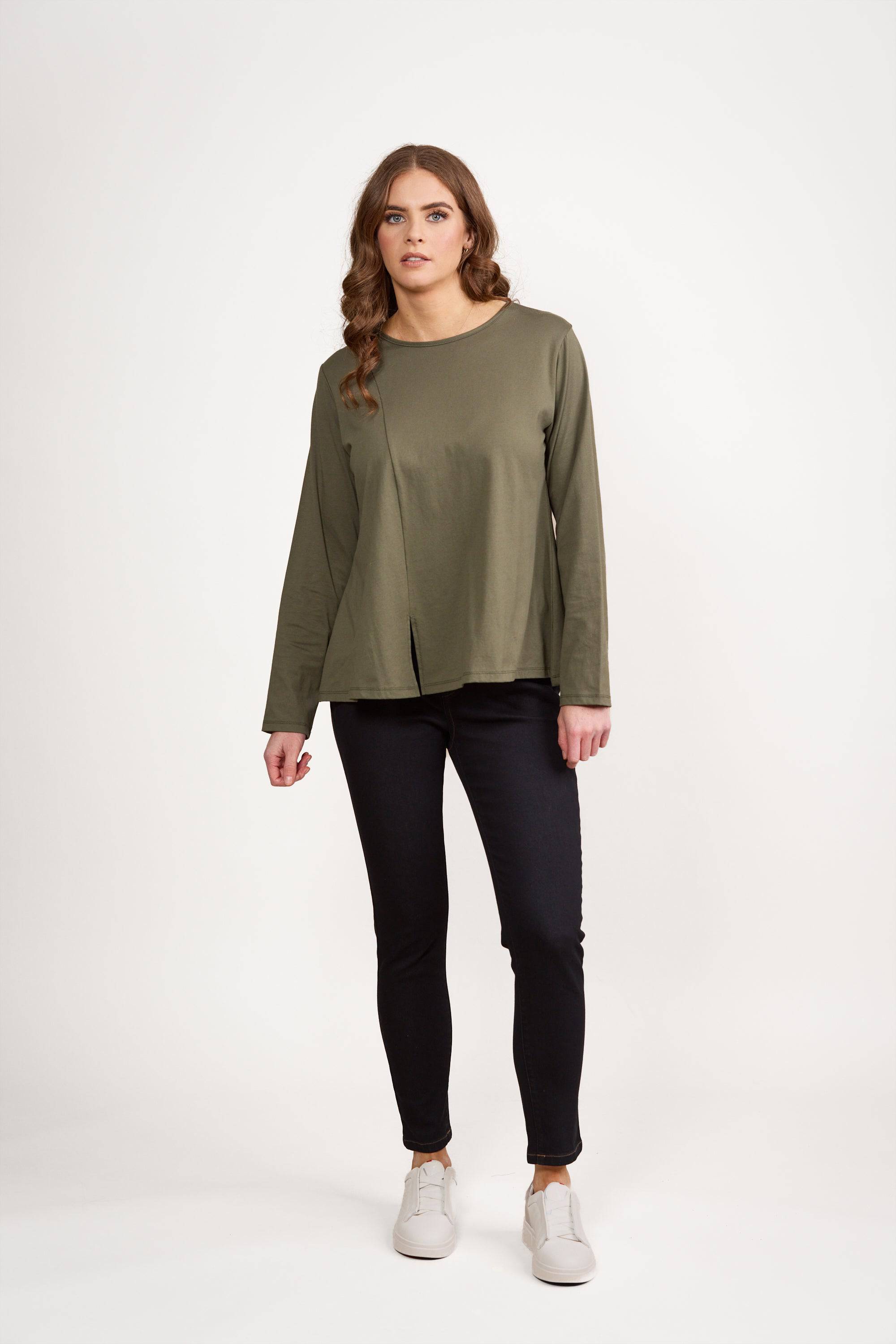 A woman with long wavy hair stands against a white background, modeling the Vassalli Swing Top in Sage. The relaxed-fit knit cotton top is paired with black pants and white sneakers as she faces forward with a neutral expression.