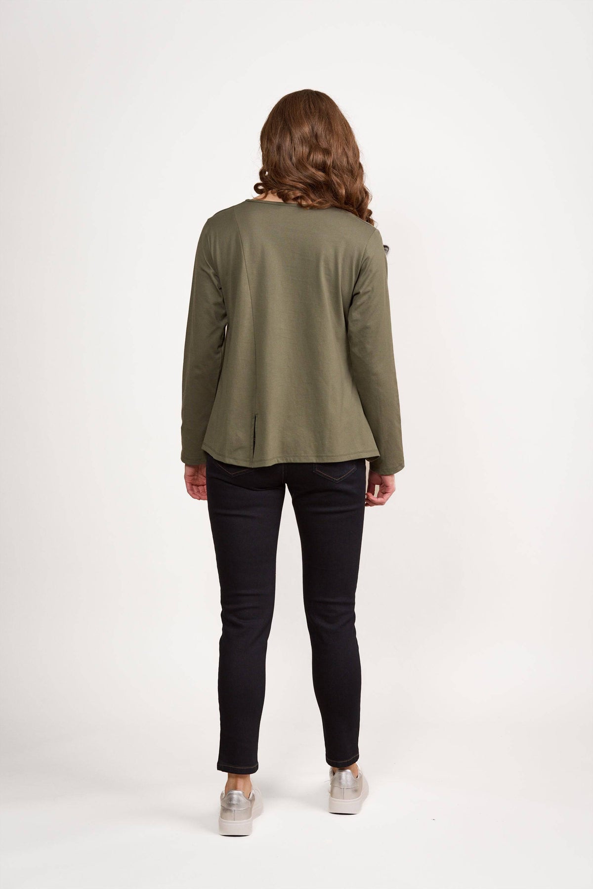 A person with wavy brown hair stands facing away on a white background, wearing the Vassalli Swing Top in Sage, styled with black pants and light sneakers.