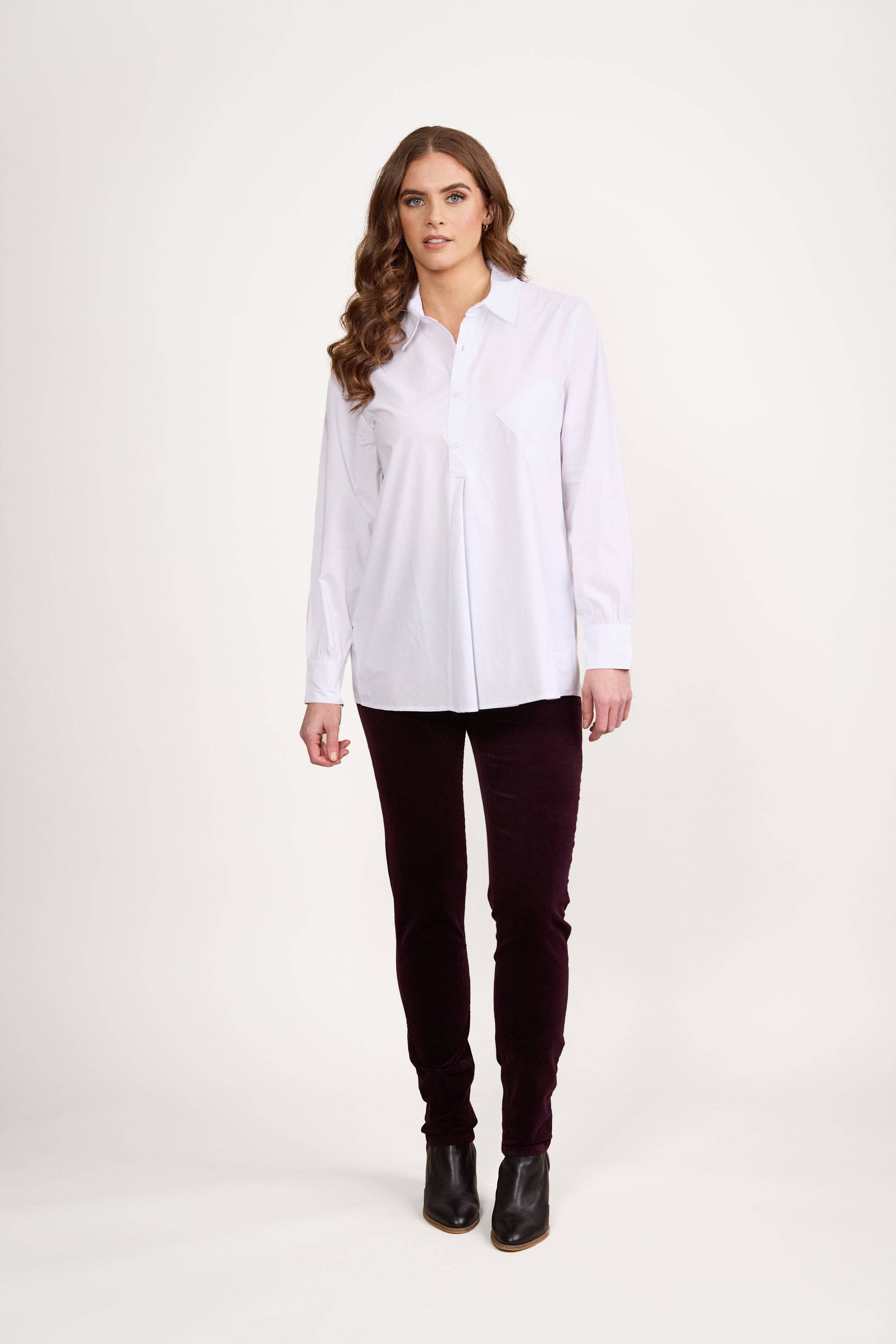 A woman stands against a plain white background, wearing the Vassalli Half Placket Shirt in white with dark pants and black ankle boots. She has long, wavy brown hair and a neutral expression.