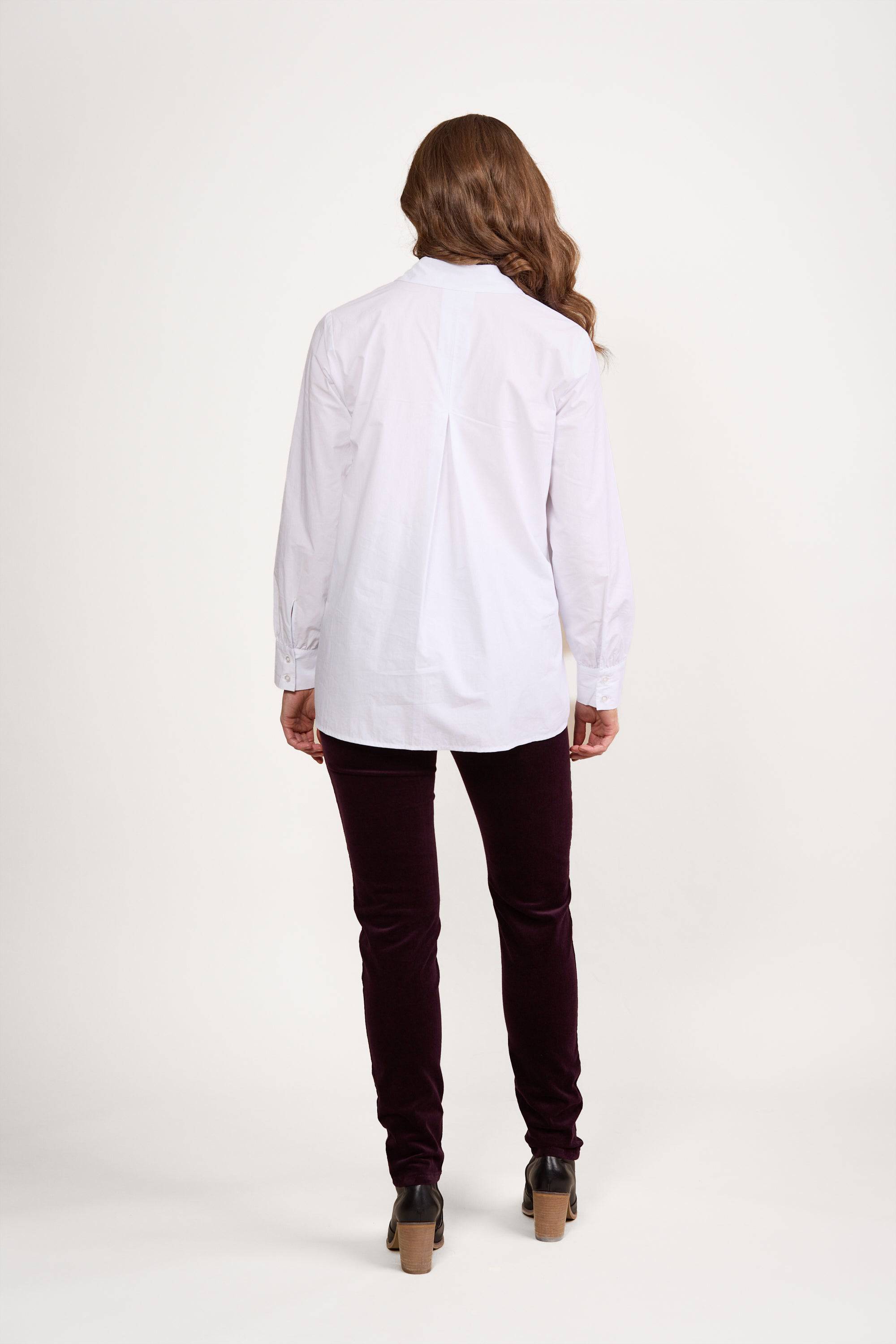 A person with long, wavy hair, wearing a relaxed fit Vassalli Half Placket Shirt in white and dark pants, stands facing away from the camera against a plain white background.