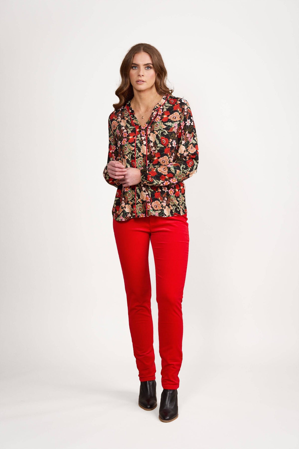 A woman with long wavy brown hair and a neutral expression stands against a white background wearing the Vassalli V-Neck Top – Flamenco, styled with bright red pants and black ankle boots.