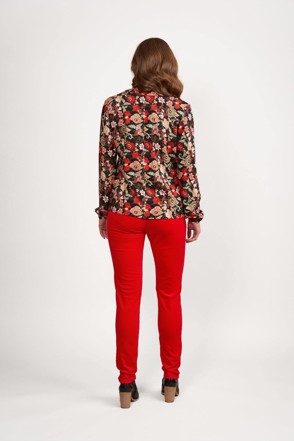 A person with wavy brown hair stands facing away, wearing the Vassalli V-Neck Top - Flamenco in red, pink, and green hues, paired with bright red pants and brown heeled shoes against a plain white background.