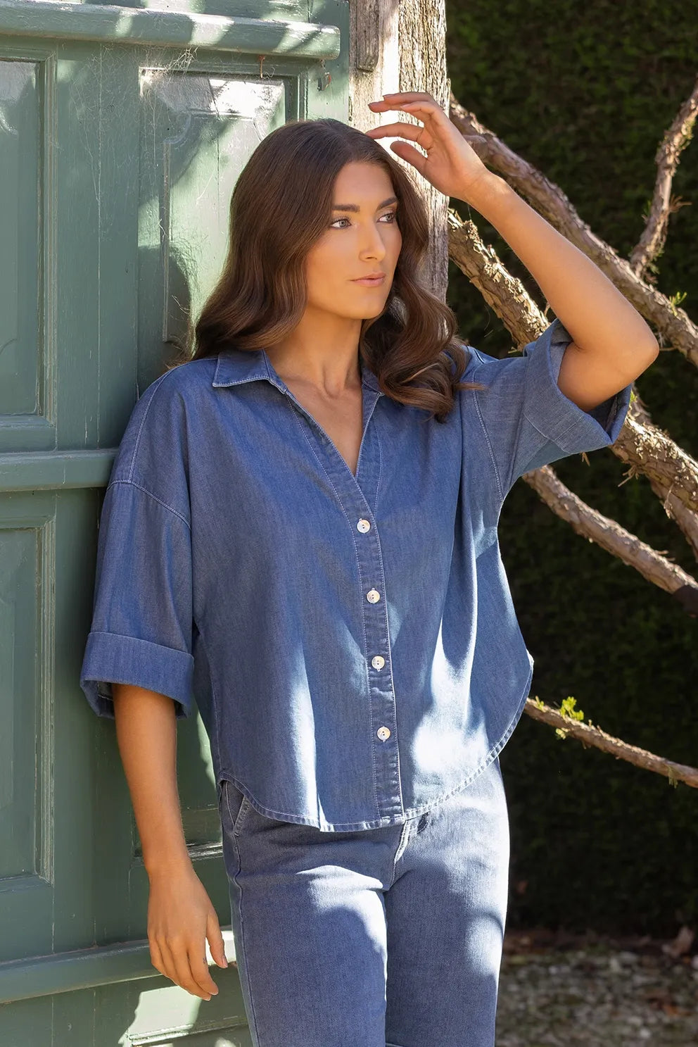 A woman with long brown hair stands outdoors by a green door, touching a wooden post and gazing into the distance. She is wearing the Vassalli Atlas Shirt in Surf Blue paired with jeans, as sunlight highlights her face and hair.