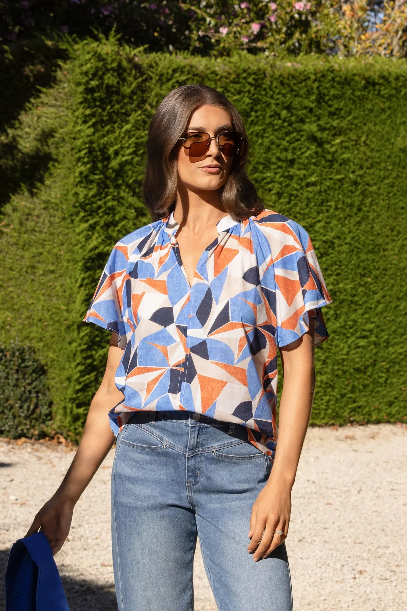 A woman stands outdoors in sunlight on a gravel path with a green hedge in the background, wearing sunglasses, light blue jeans, and the Vassalli Nyla Top - Row—a colorful geometric print cotton blouse with flutter sleeves.