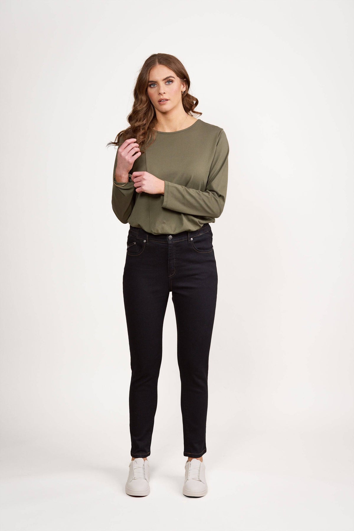 A woman with long wavy brown hair stands against a white background, wearing an olive green long-sleeve shirt, Vassalli Mode Jean in black denim, and white sneakers. She is holding a lock of her hair with one hand.
