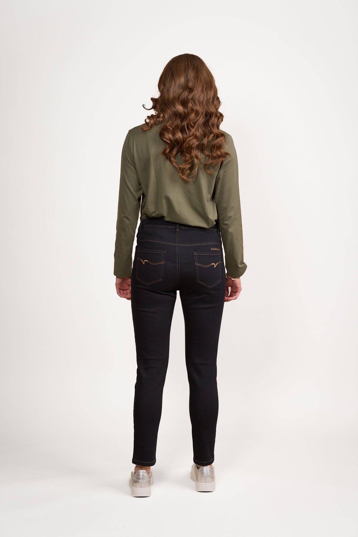 A person with long, wavy brown hair stands facing away, wearing a green long-sleeve shirt, Vassalli Mode Jean in black denim crafted from stretch fabric for a comfortable fit, and light-colored shoes, against a plain white background.