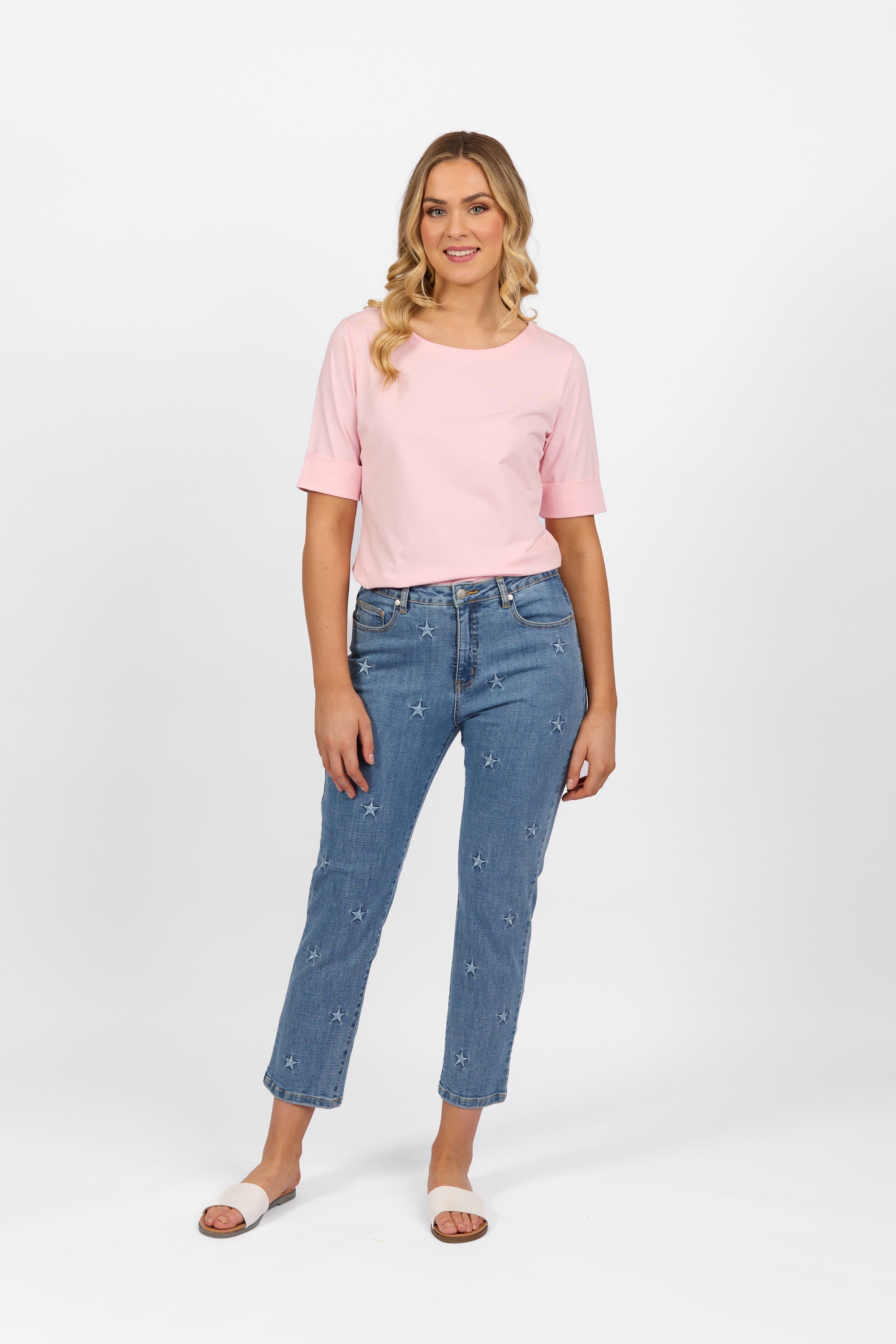 A woman smiles against a plain white background, wearing a light pink T-shirt, Vassalli Onya Jean - Light Wash, and white slide sandals.