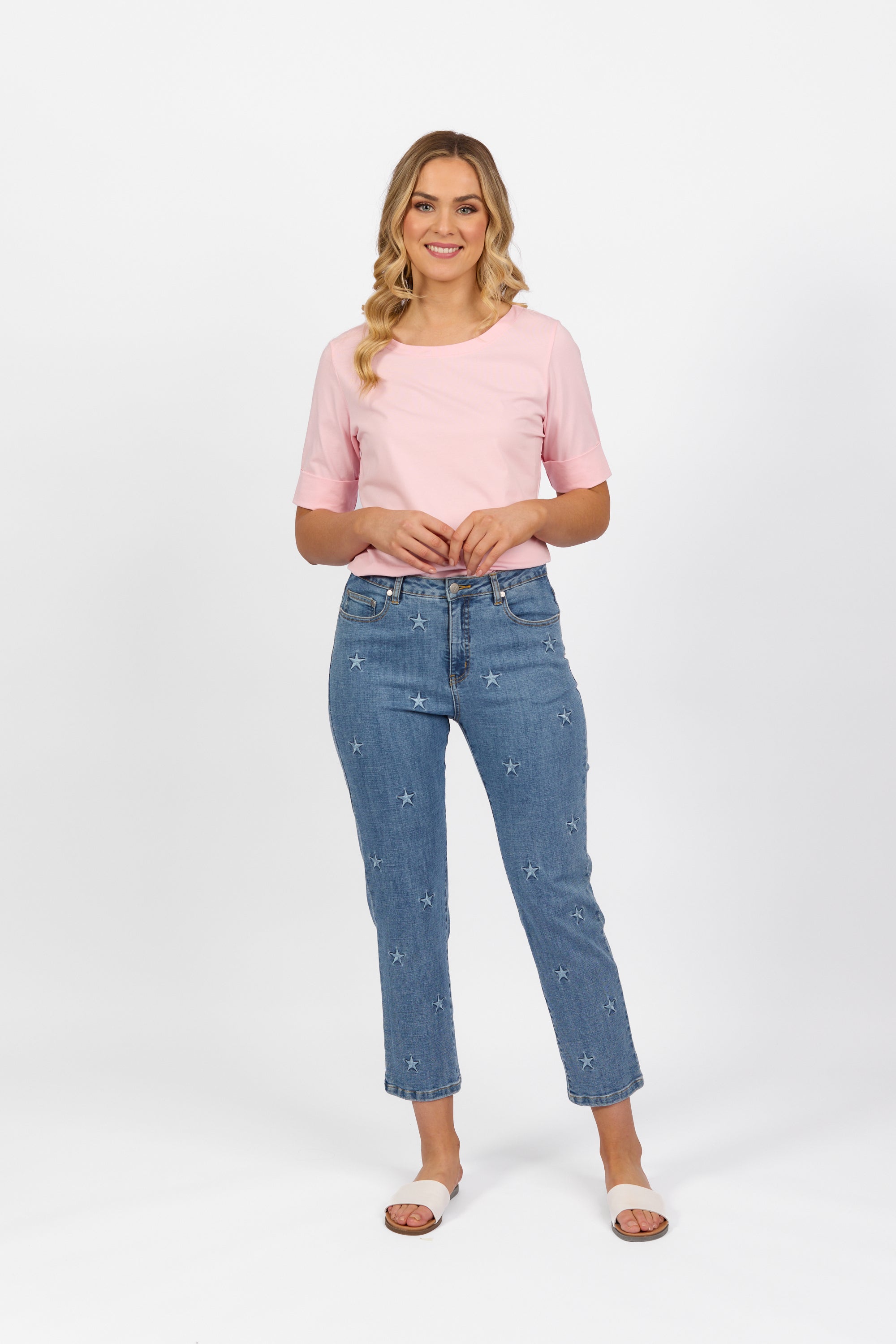A woman with long wavy hair smiles against a white background, wearing a light pink short-sleeve top, Vassalli Onya Jean - Light Wash, and white sandals.