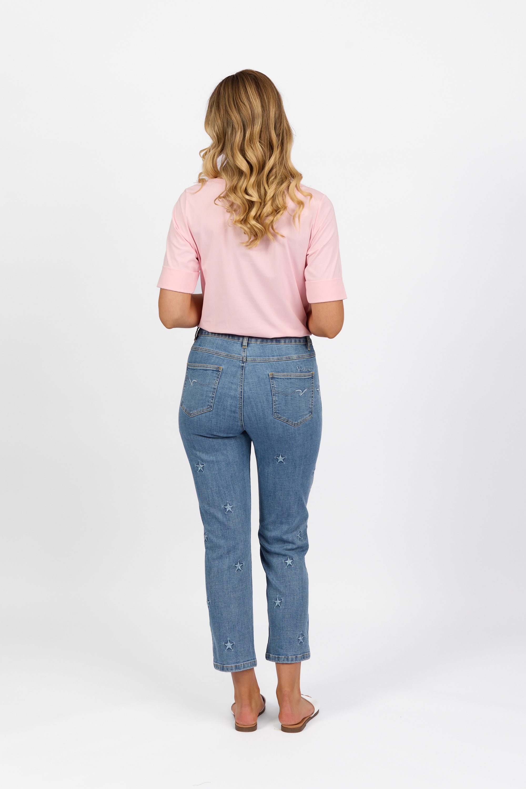 A woman with long wavy blonde hair wears a light pink short-sleeve shirt and Vassalli Onya Jean - Light Wash, standing in sandals facing away from the camera against a plain white background.
