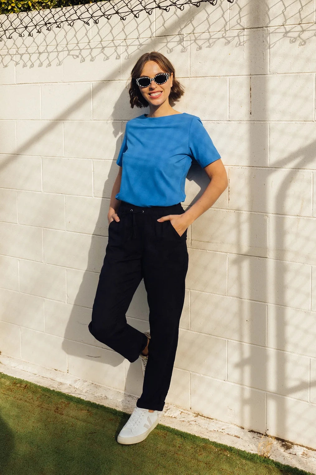 A woman in the Vassalli Oxford Pant - Ink, paired with a blue t-shirt and white sneakers, leans against a white brick wall with her hands in her pockets. She wears oversized patterned sunglasses and smiles, her short hair and relaxed pose highlighting the effortless comfort of these casual summer pants crafted from a soft linen-viscose blend.