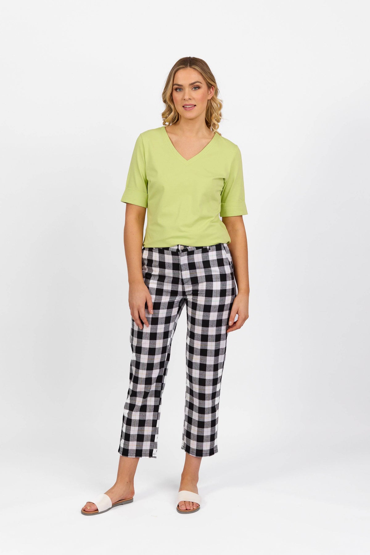 A woman with light wavy hair stands against a white background, wearing a light green V-neck short-sleeve top, Vassalli 7/8 Pant - Rainbow Check in black and white, and white slide sandals, looking at the camera.