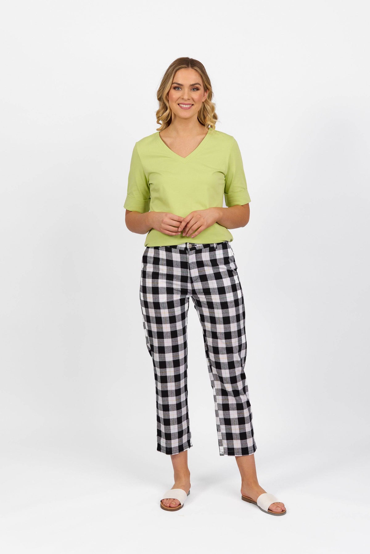 A woman stands smiling, wearing a light green short-sleeve top, Vassalli 7/8 Pant - Rainbow Check, and white open-toe sandals against a plain white background.
