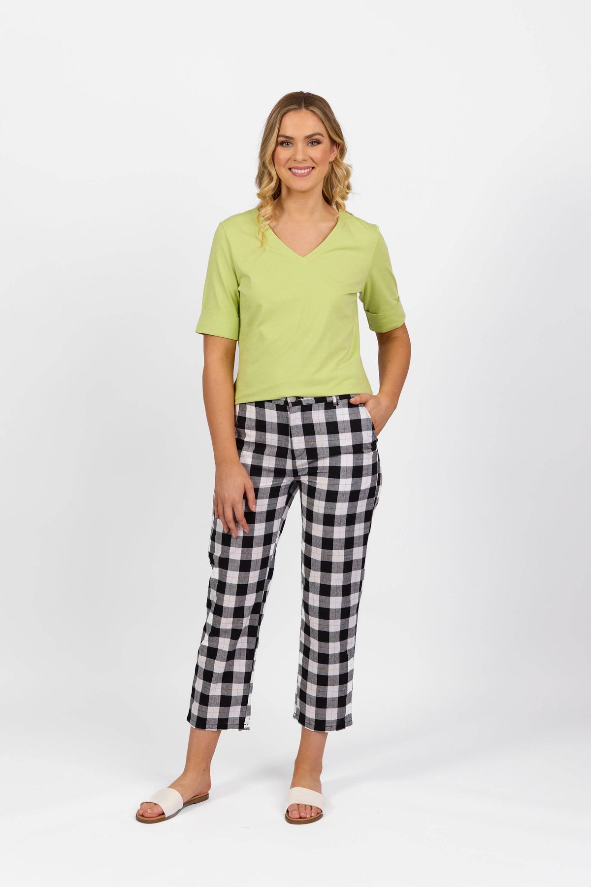 A woman stands against a white background wearing a light green top, Vassalli 7/8 Pant - Rainbow Check, and white slide sandals. She is smiling with one hand in her pocket.