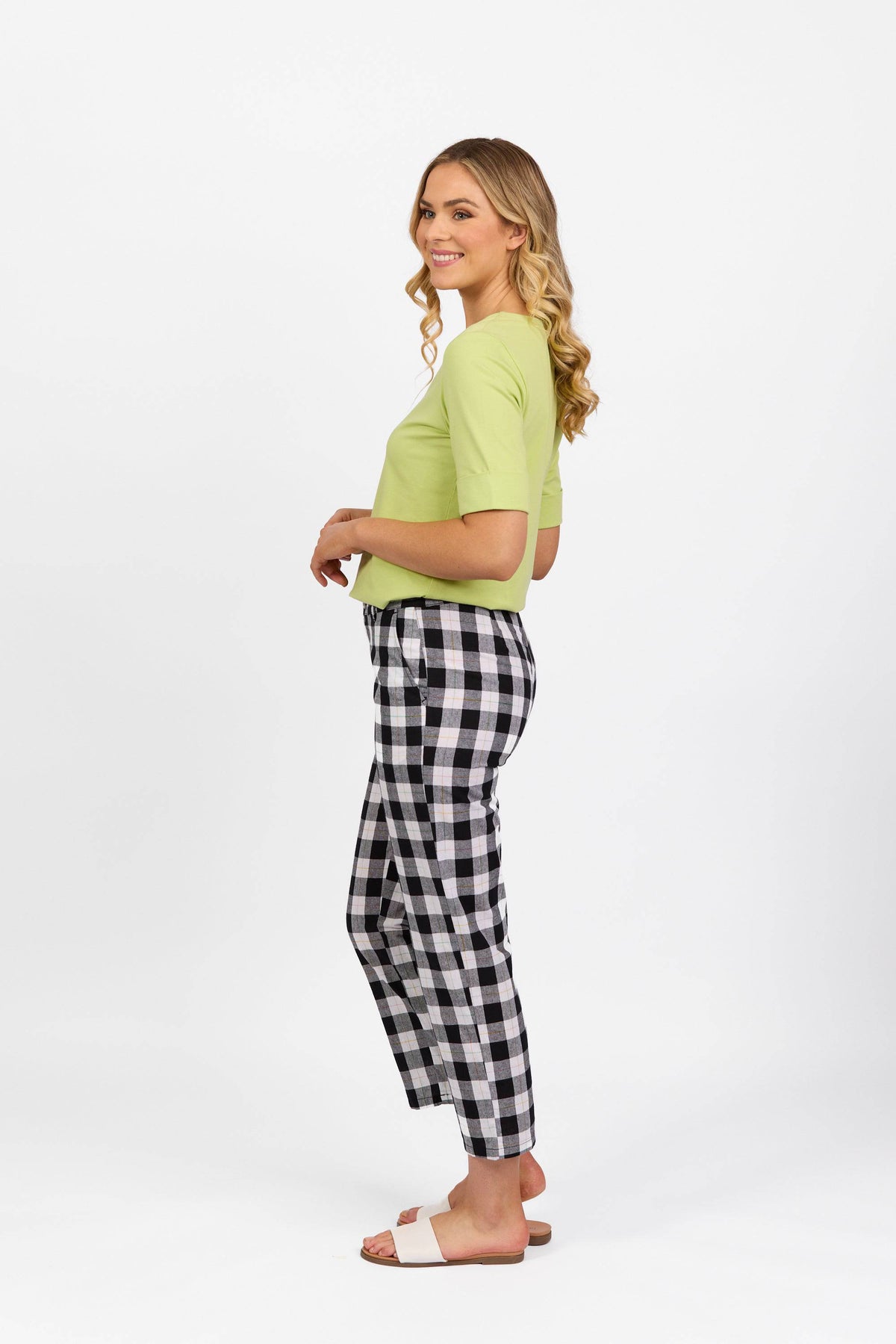 A smiling woman with long blonde hair stands sideways, wearing a light green top, Vassalli 7/8 Pant - Rainbow Check, and white sandals against a plain white background.