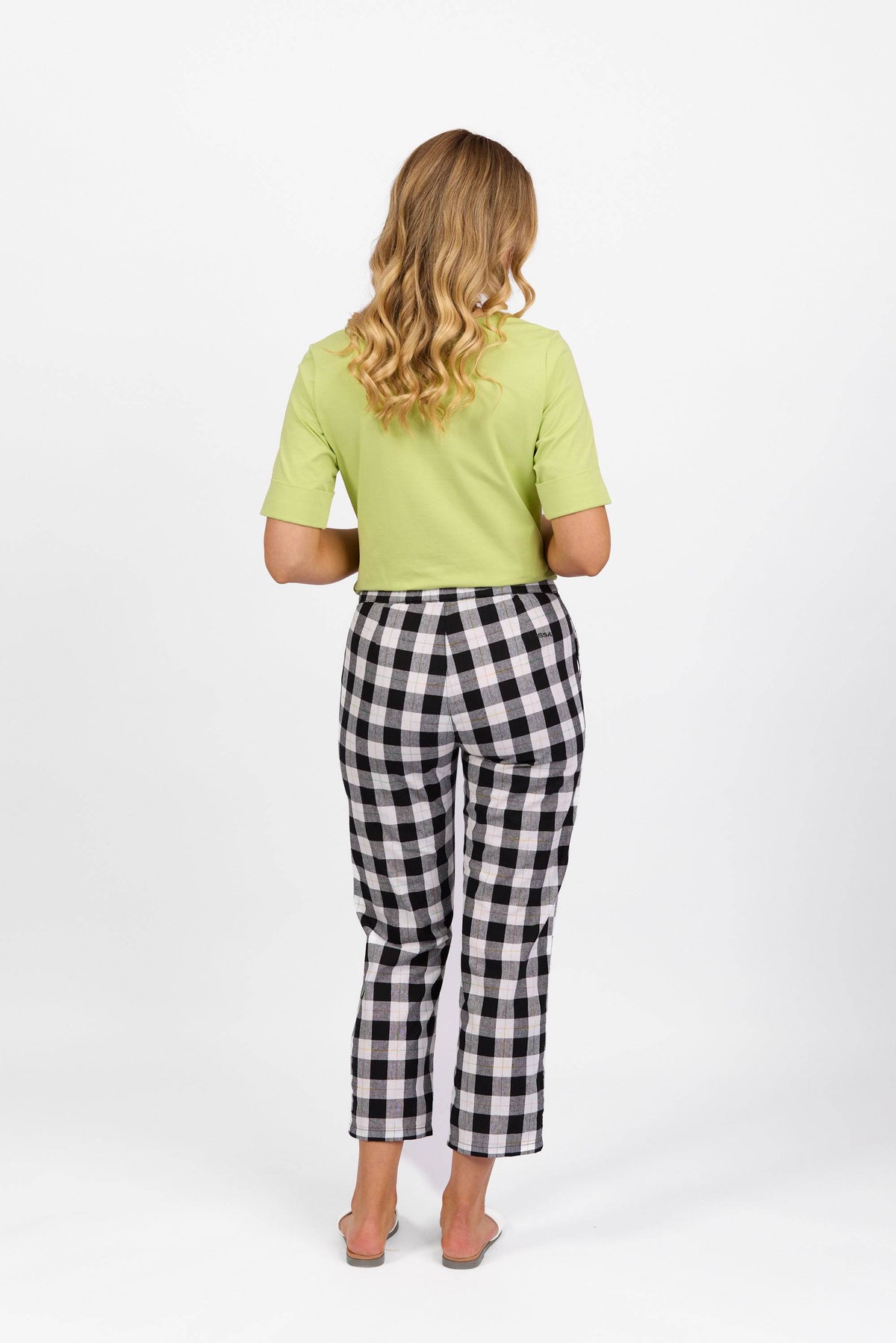 A woman with wavy blonde hair, barefoot against a plain white background, wears a light green short-sleeve top and Vassalli's 7/8 Pant - Rainbow Check, featuring a high waist and cropped slim legs.