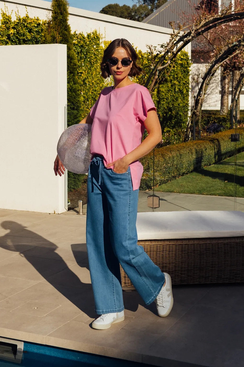 A woman stands poolside on a sunny day, smiling as she holds a sunhat. She wears sunglasses, a pink top, and the Vassalli Yarra Jean - Mid Denim: stylish blue wide-leg jeans crafted from stretch denim. Trees, bushes, and white walls create a serene background.