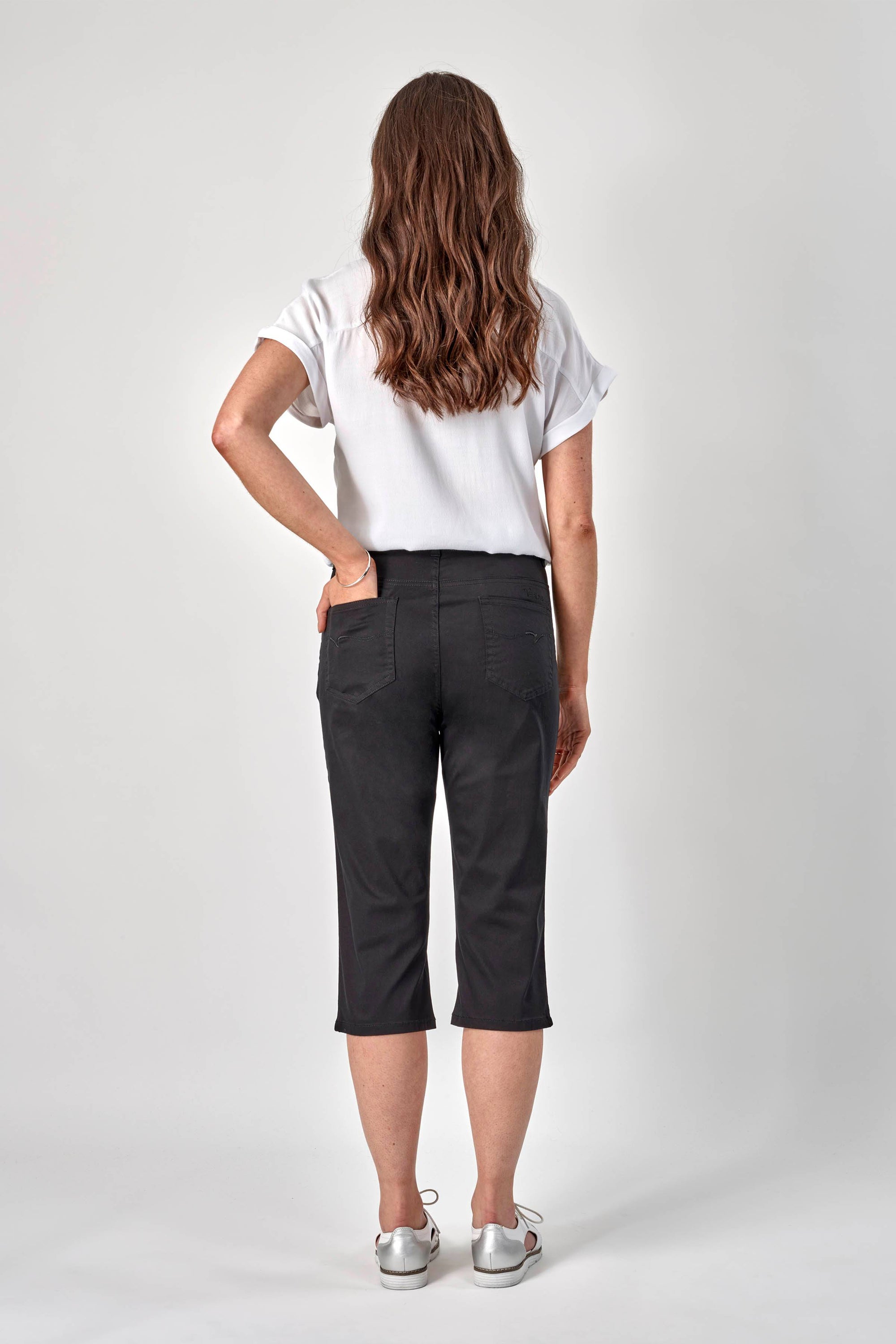 A woman with long wavy brown hair, wearing a white short-sleeve blouse, Vassalli 3/4 Pant - Black, and white sneakers, stands facing away from the camera against a plain light background.