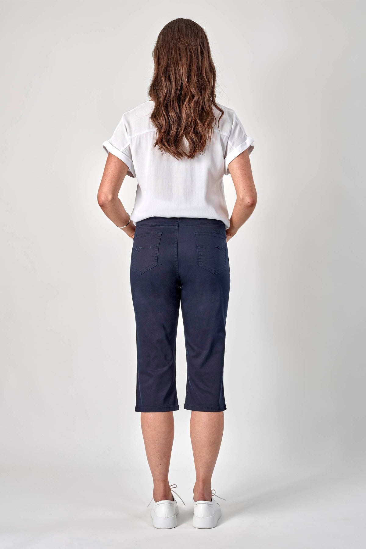 A woman with long wavy brown hair faces away, wearing a white short-sleeve shirt, Vassalli 3/4 Pant in navy, and white sneakers against a plain light gray background.