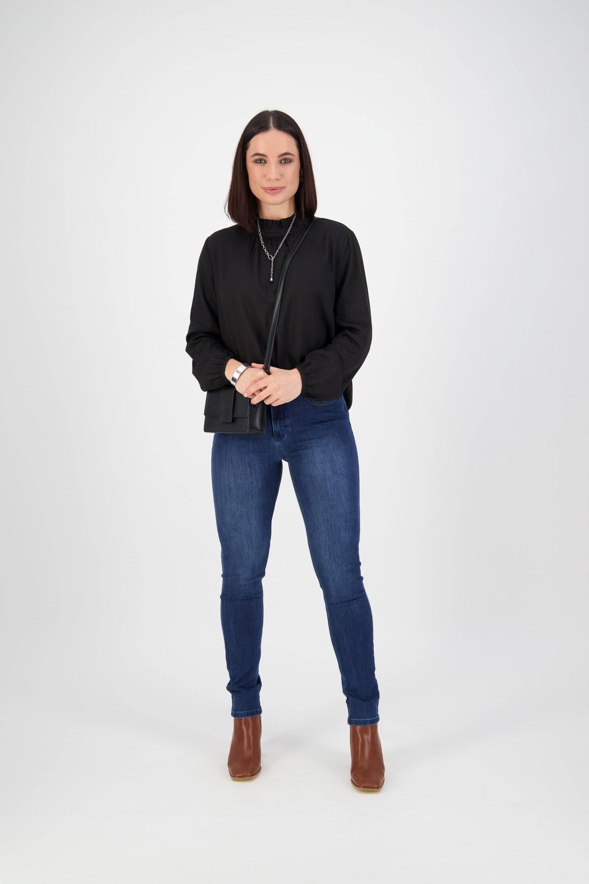 A woman with straight, shoulder-length dark hair faces the camera in a black top, Vassalli Skinny Leg Jean - Blue Denim, brown ankle boots, and a necklace, set against a plain white background.