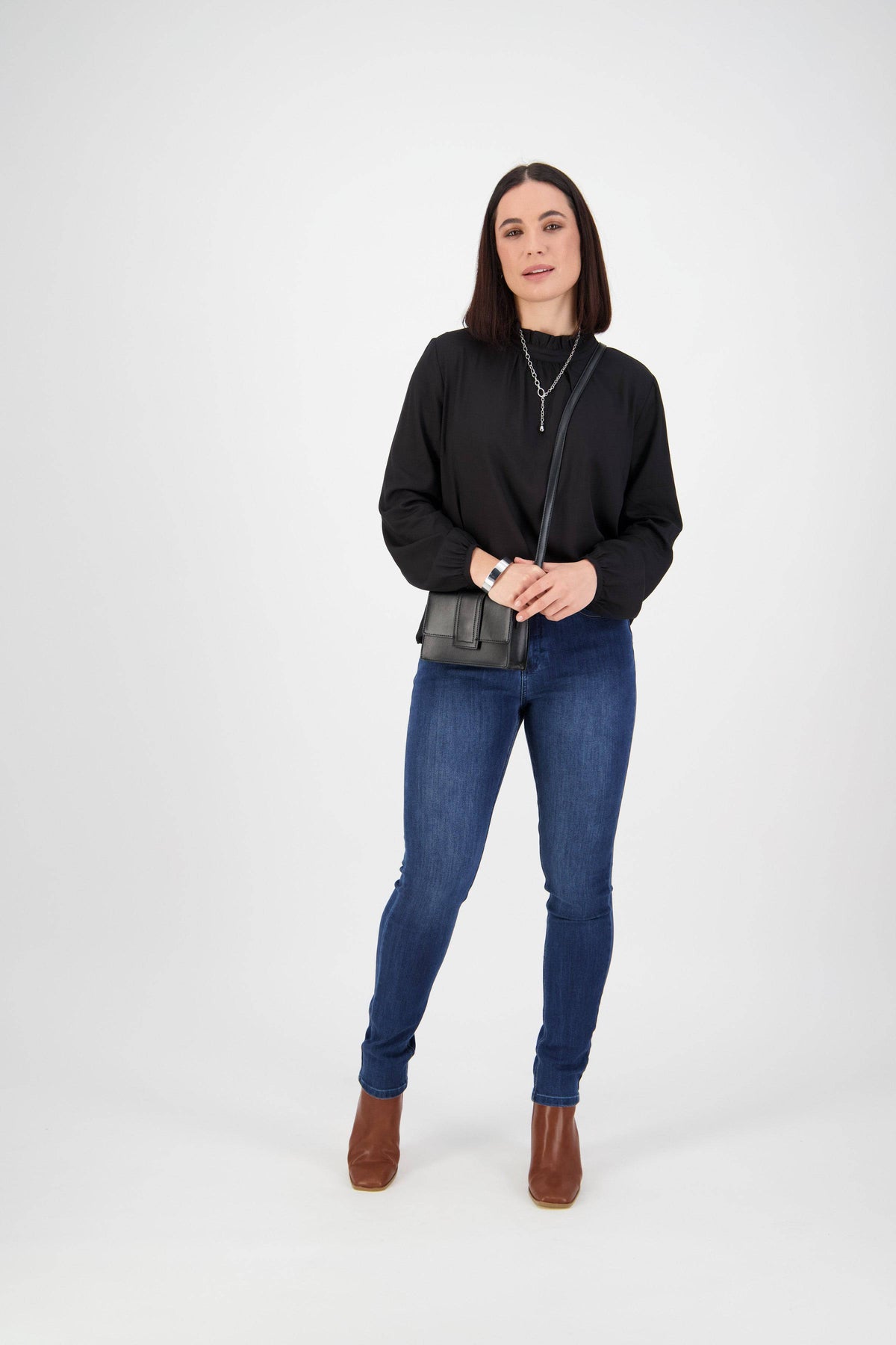 A woman stands against a white background wearing a black long-sleeve top, Vassalli Skinny Leg Jean in Blue Denim, brown ankle boots, and a small black crossbody purse. She has straight dark hair and looks slightly to the side.