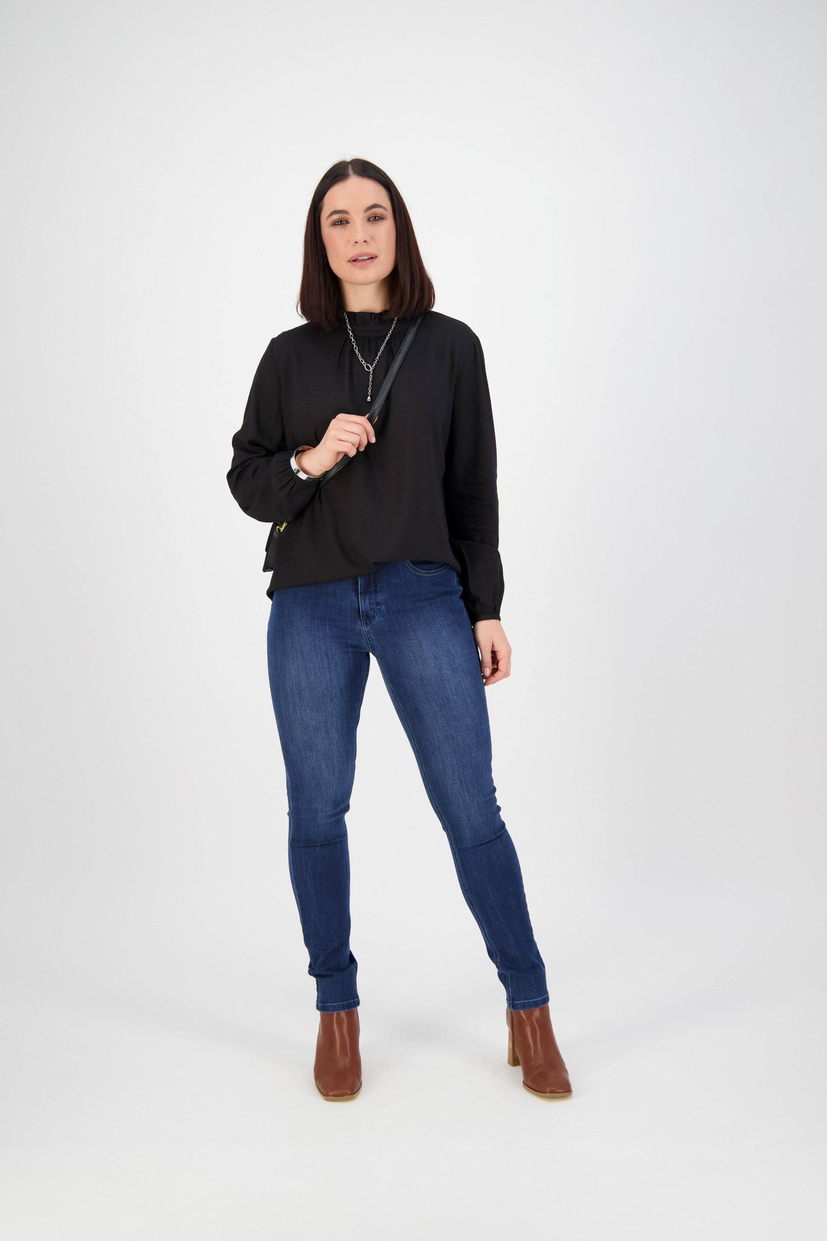 A woman with straight dark hair wears a black long-sleeve top, Vassalli Skinny Leg Jean in Blue Denim, brown ankle boots, and layered necklaces, standing against a plain white background.
