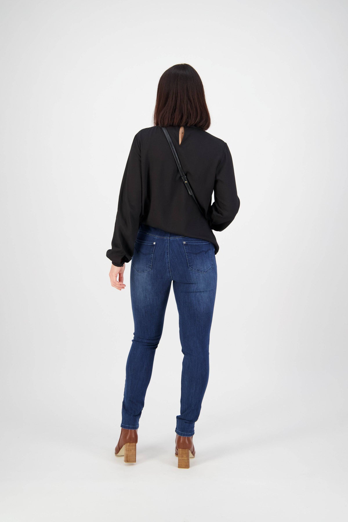 A person with shoulder-length brown hair, wearing a black long-sleeve top and Vassalli Skinny Leg Jean - Blue Denim, stands facing away from the camera against a plain light background.