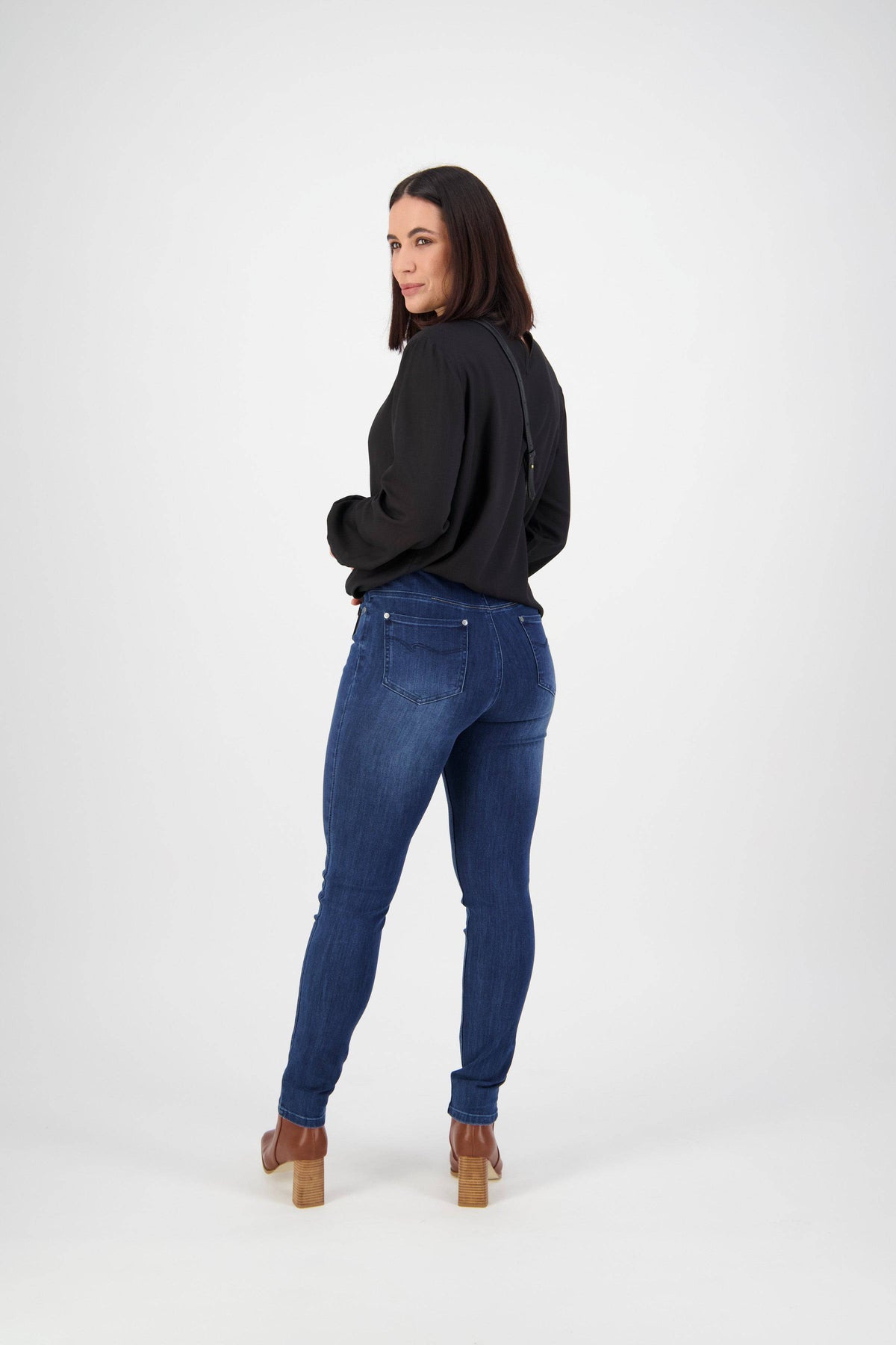 A woman with shoulder-length dark hair poses against a plain white background, looking over her shoulder. She wears a black long-sleeve top and Vassalli Skinny Leg Jean - Blue Denim with tan heeled shoes.