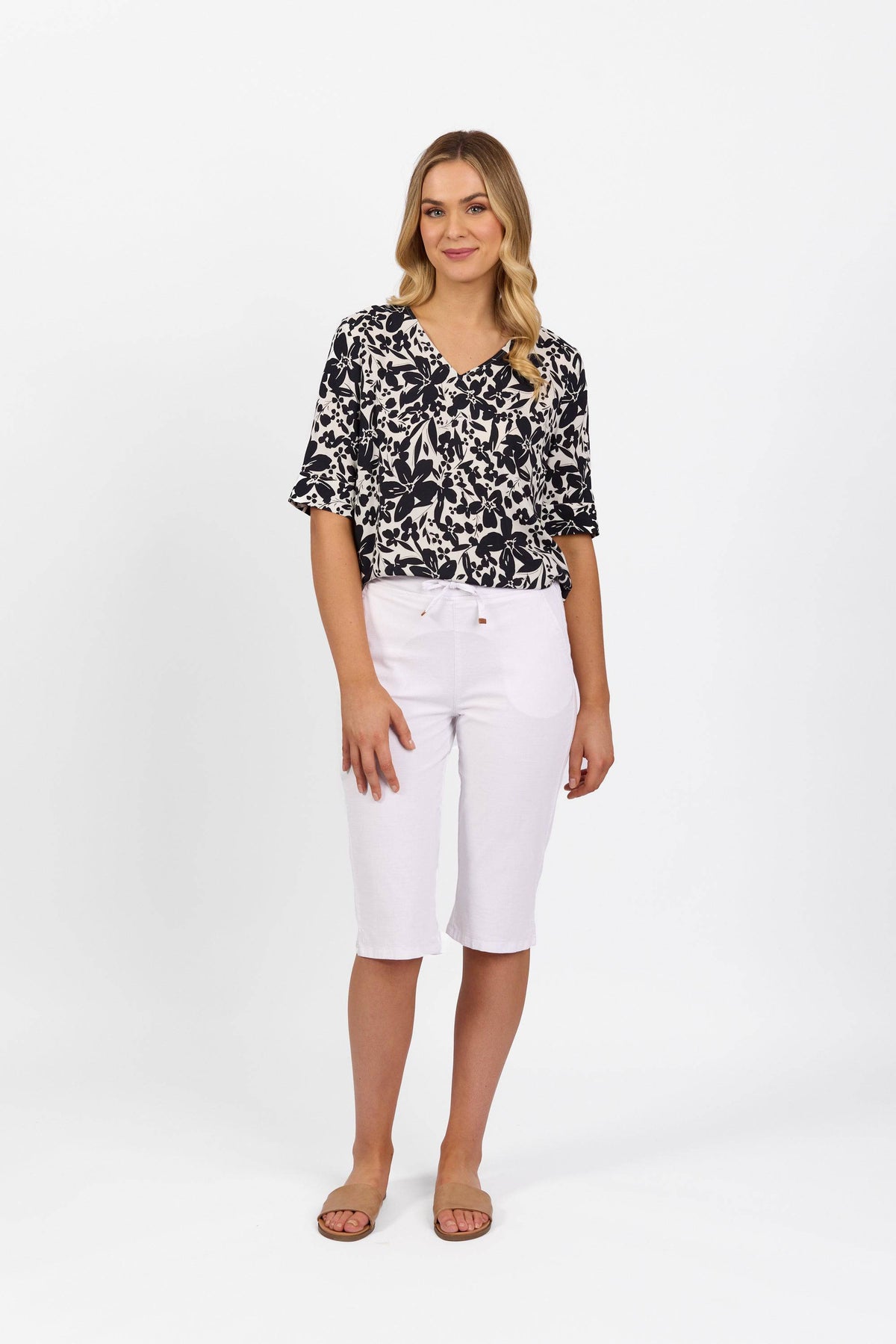 A woman with long, wavy blonde hair stands against a white background, smiling slightly. She wears a black and white patterned blouse, tan sandals, and Vassalli's Drew 3/4 Pant in white—knee-length stretch cotton shorts with an elastic waistband.