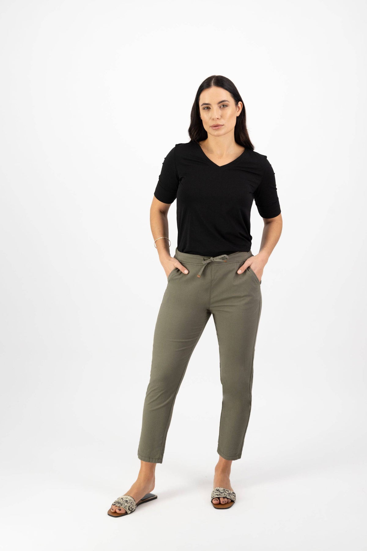 A woman with long dark hair and a neutral expression stands against a white background wearing Vassalli Denver 7/8 Pant in Army, paired with a black V-neck top and patterned slide sandals, her hands tucked into her pockets.