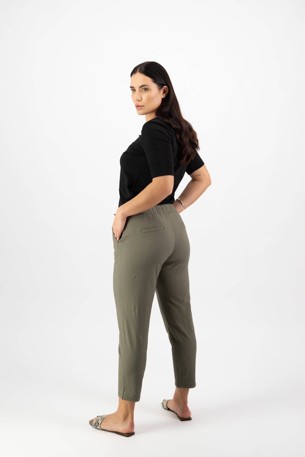 A woman with long dark hair stands in a three-quarter pose, wearing the Vassalli Denver 7/8 Pant in Army and a black top, paired with flat sandals, against a plain white background.