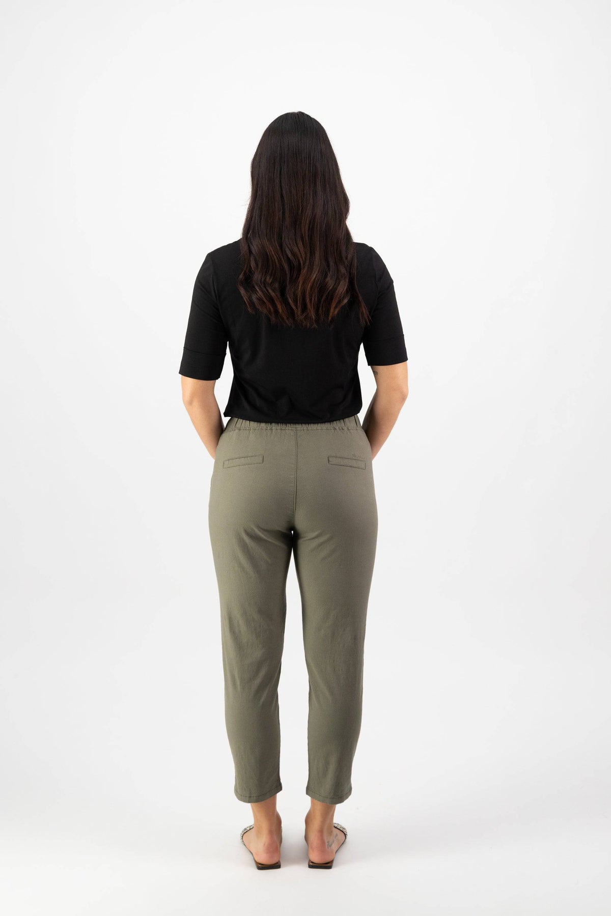A woman faces away from the camera, barefoot against a white background, wearing the Vassalli Denver 7/8 Pant in army green and a black short-sleeve top.