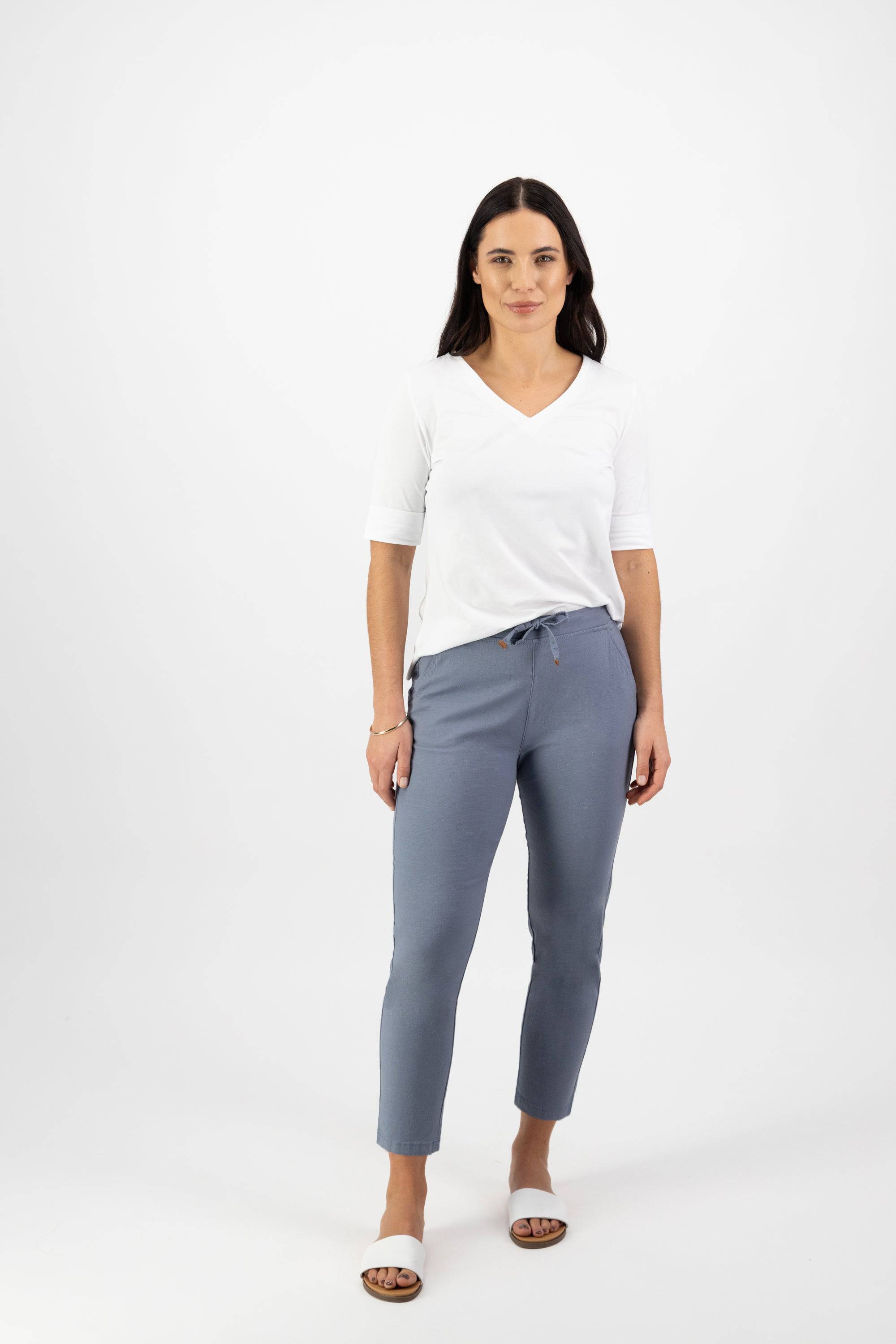 A woman with long dark hair stands against a white background, wearing a white V-neck top and Vassalli's Denver 7/8 Pant in Atlantic. The light blue pants have an elastic waist and skinny leg. She pairs them with brown sandals, hands relaxed by her sides.