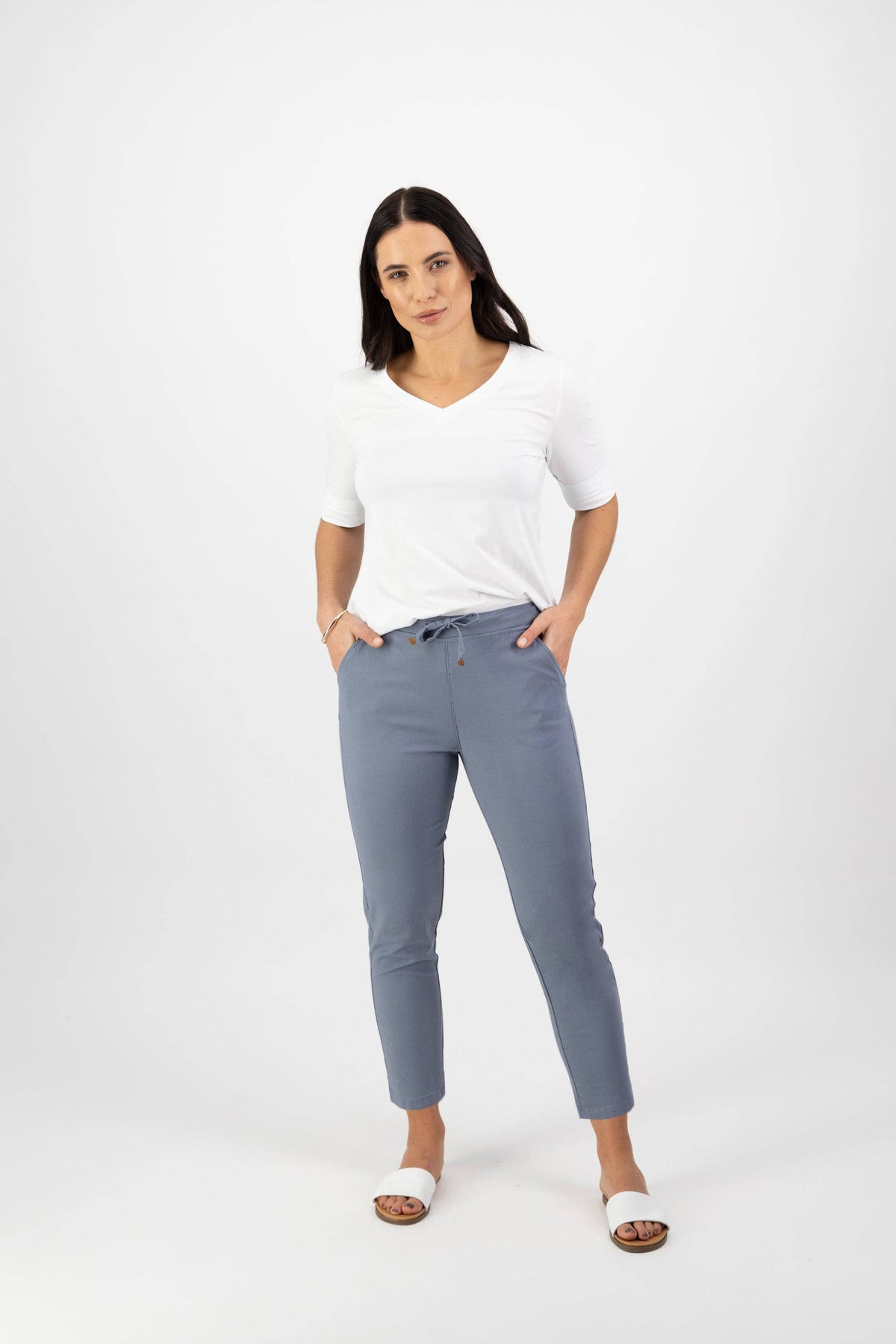 A woman with long dark hair stands against a white background, wearing a white V-neck T-shirt and Vassalli Denver 7/8 Pant in Atlantic. The stretch fabric pants have a skinny leg and elastic waist; she stands with hands in her pockets and a neutral expression.