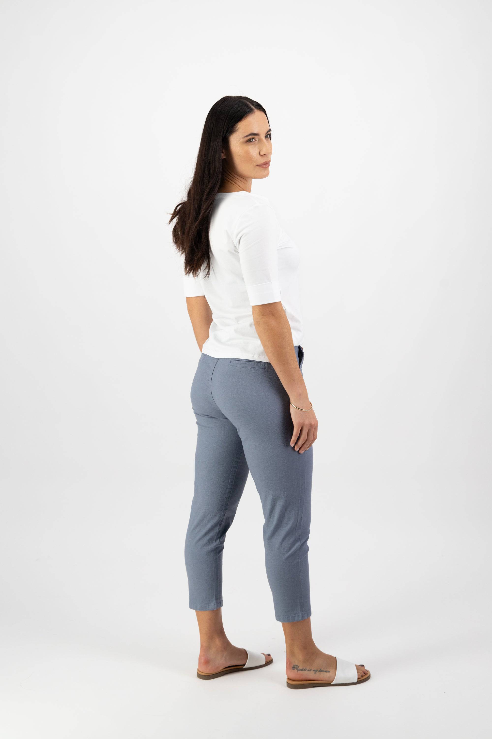 A woman with long brown hair stands in a white room, glancing over her shoulder. She wears a white top, Vassalli Denver 7/8 Pant - Atlantic, made from stretch fabric, and white slide sandals.