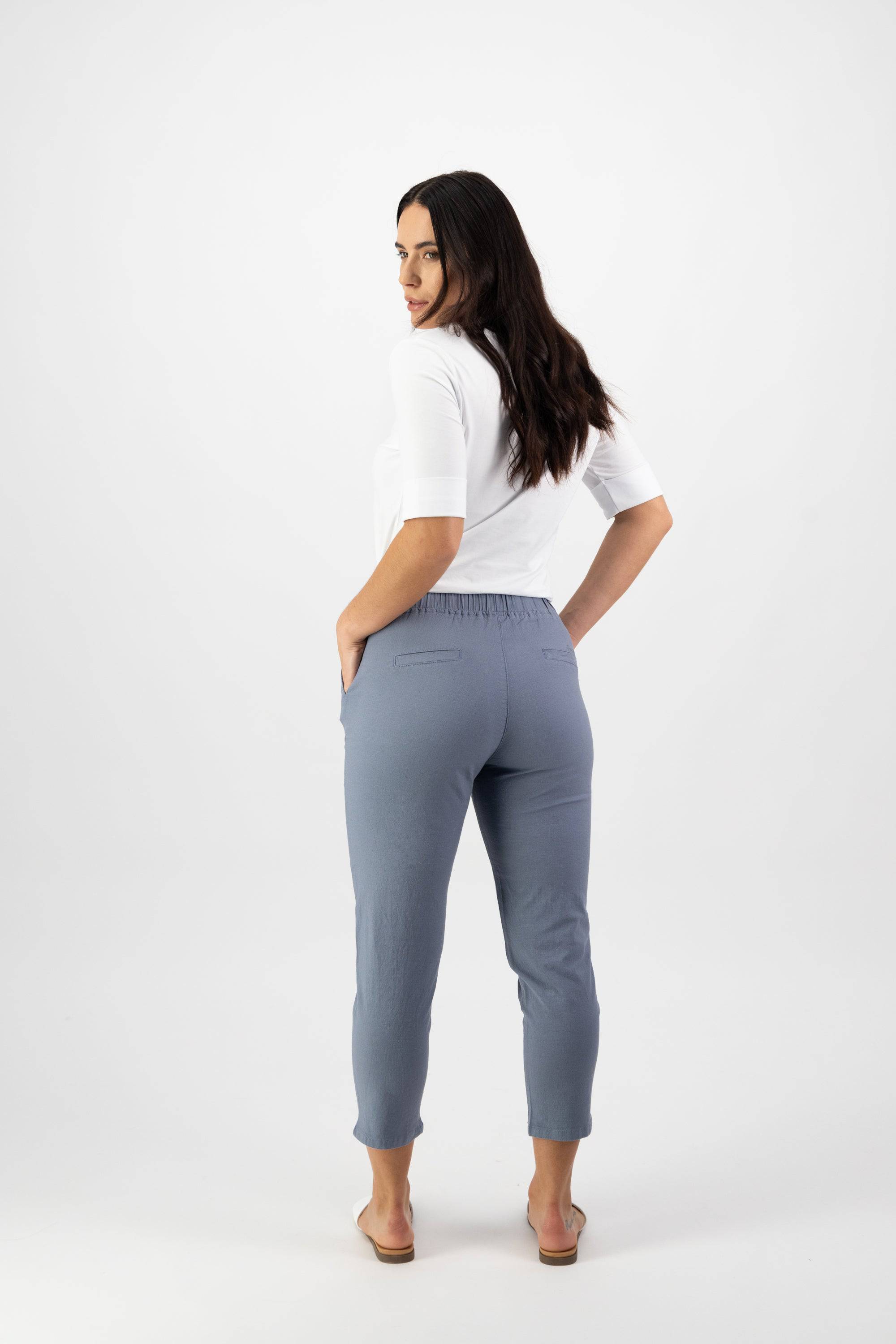 A woman with long dark hair, facing away and looking over her shoulder, wears a white top, Vassalli Denver 7/8 Pant in Atlantic, and beige sandals against a plain white background.