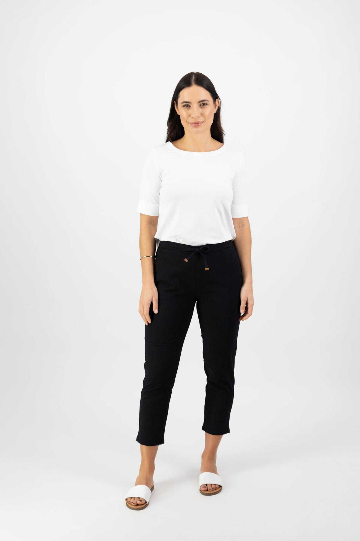 A woman stands against a plain white background wearing Vassalli's Denver 7/8 Pant in black, paired with a white short-sleeve top and white slide sandals. Her long brown hair frames her neutral expression.