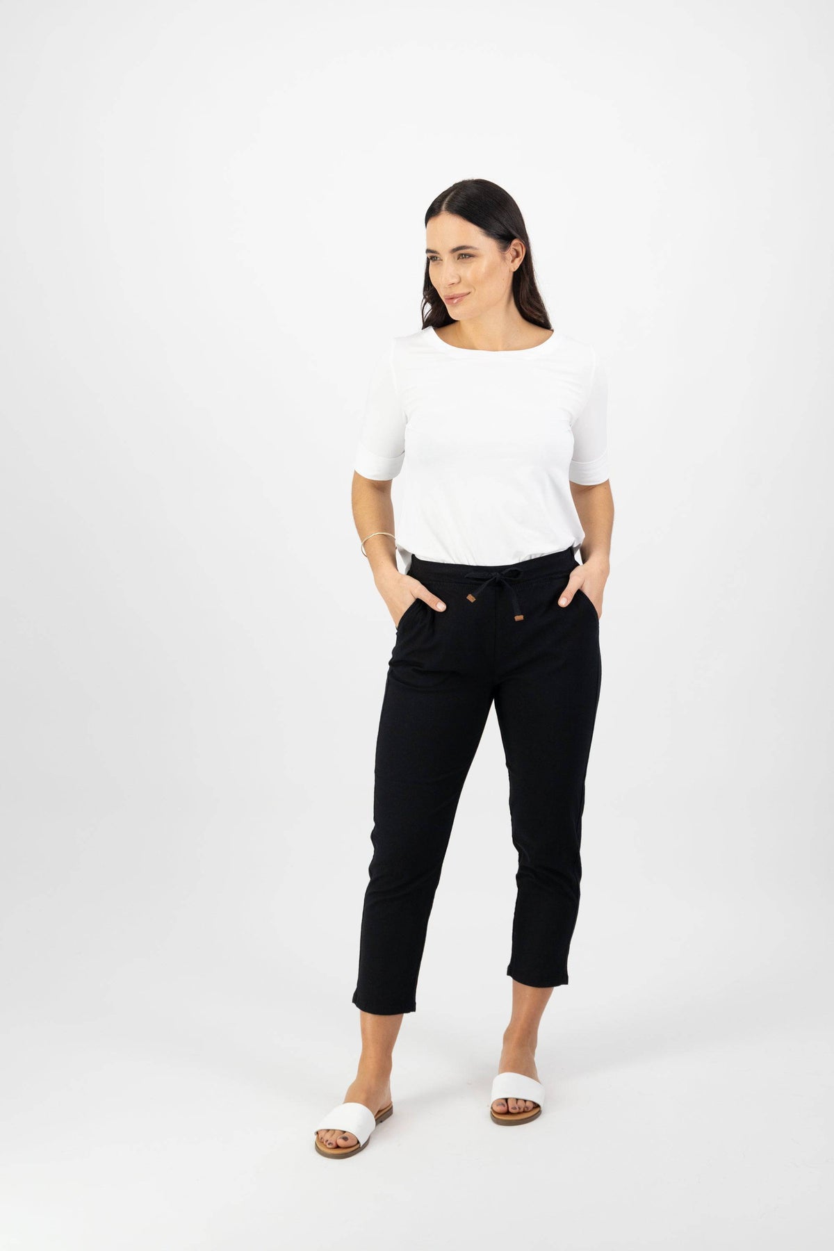 A woman stands against a white background, wearing the Vassalli Denver 7/8 Pant in black with an elastic waistband and pockets, paired with a white short-sleeve top and white slide sandals. She looks to her left with a neutral expression.