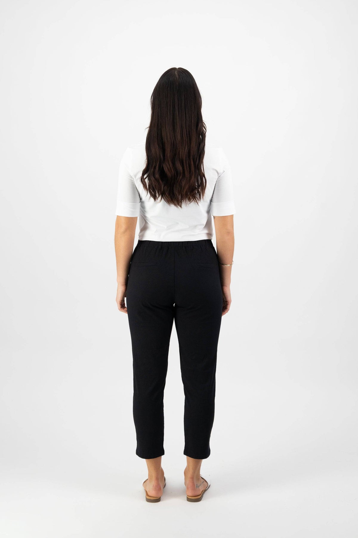 A woman with long dark hair, facing away, wears a white short-sleeve shirt and Vassalli's Denver 7/8 Pant in Black with an elastic waistband and sandals, set against a plain white background.