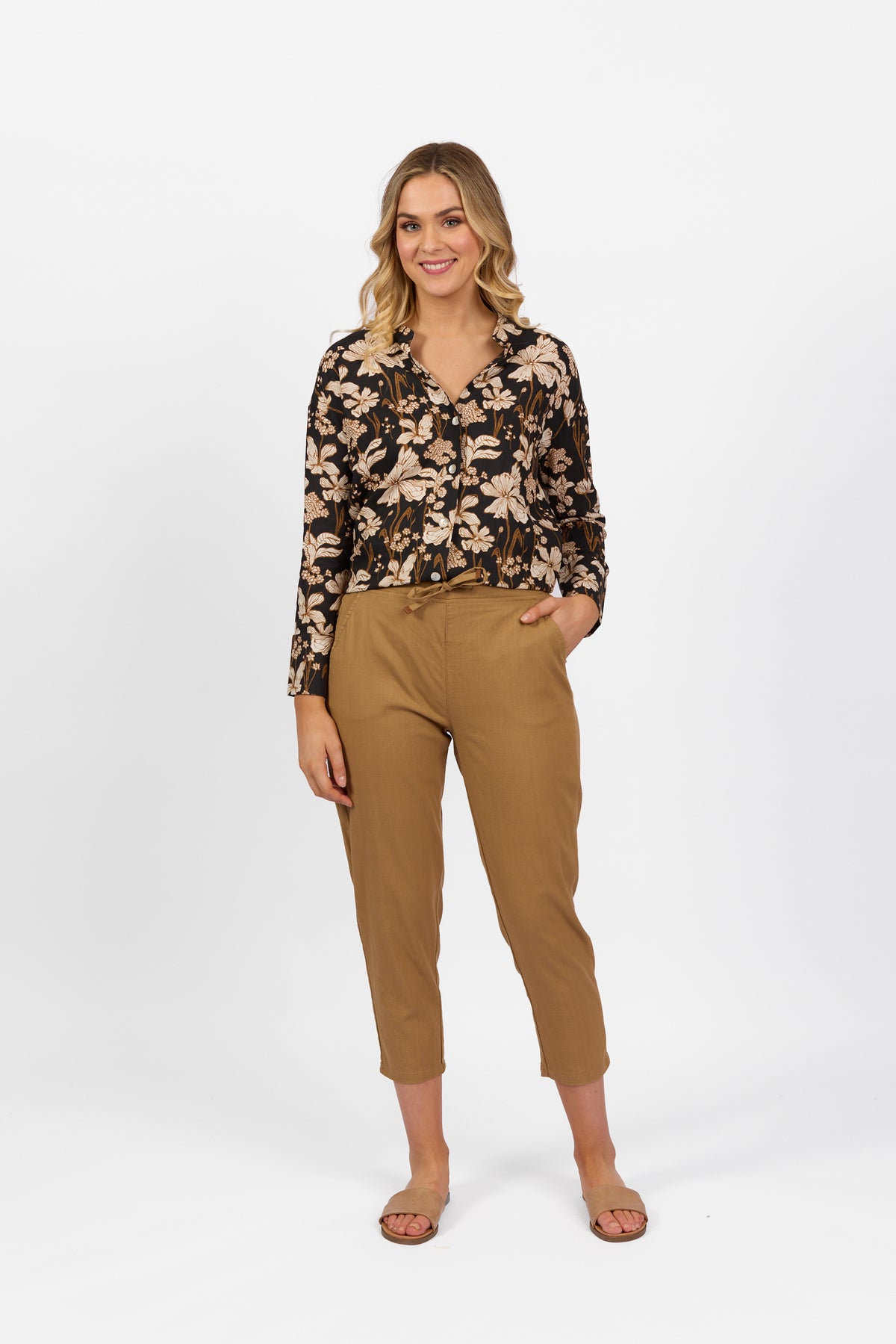 A woman stands against a white background, wearing a black floral long-sleeve blouse, Vassalli Getaway Pant in Caramel with an elastic waist and skinny leg, and tan sandals. She has wavy shoulder-length blonde hair and smiles with one hand in her pocket.