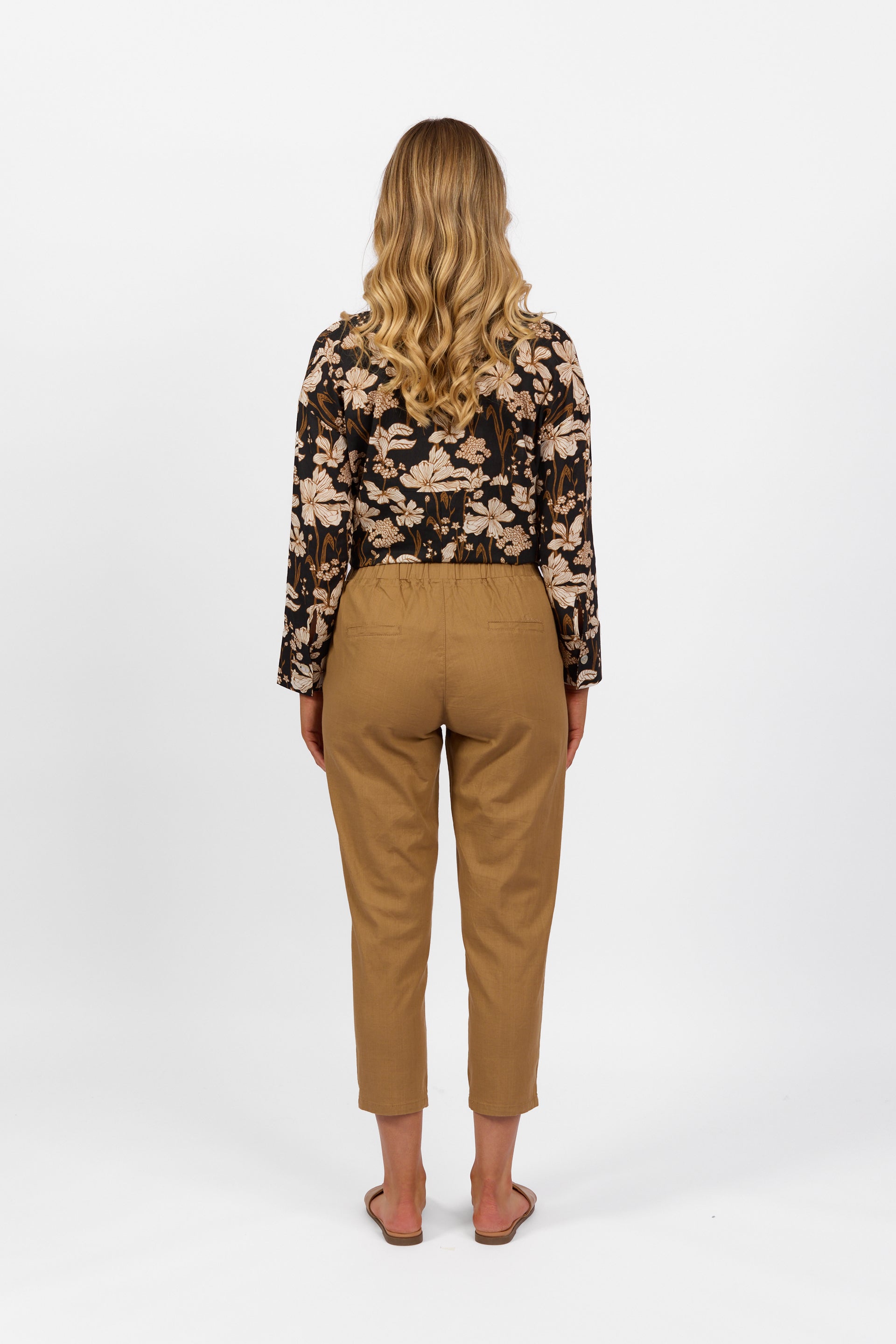 A woman with long, wavy blonde hair, facing away, wears a black floral long-sleeve top, Vassalli Getaway Pant in Caramel with an elastic waist and skinny leg, plus flat sandals, against a plain white background.