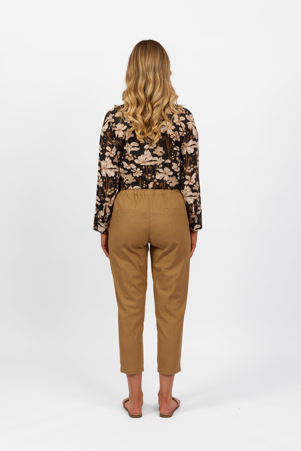 A woman with long, wavy blonde hair, facing away, wears a black floral long-sleeve top, Vassalli Getaway Pant in Caramel with an elastic waist and skinny leg, plus flat sandals, against a plain white background.